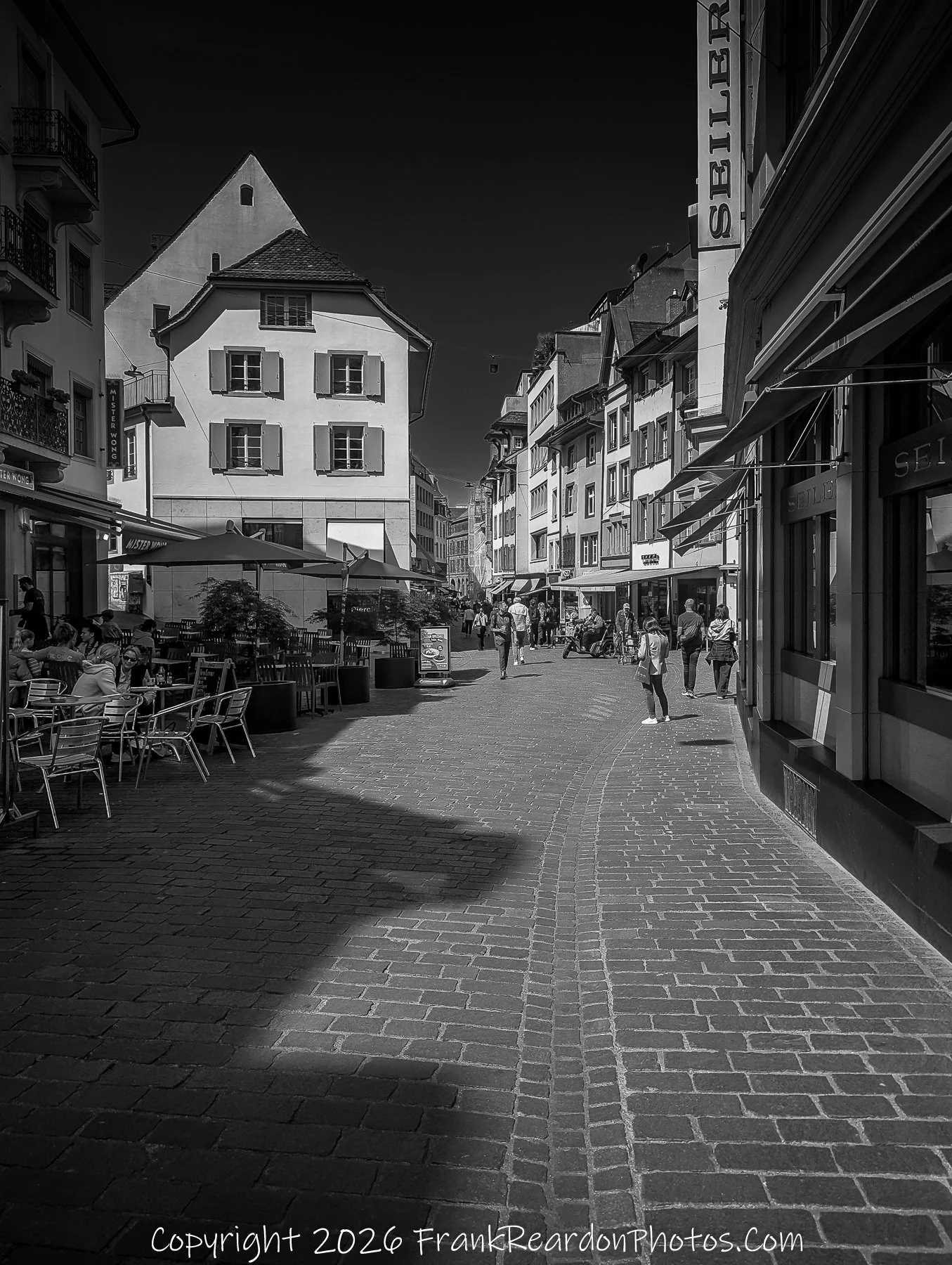 “Café Shadows” Cobblestones and sunlight weave together the rhythm of conversation and leisure.