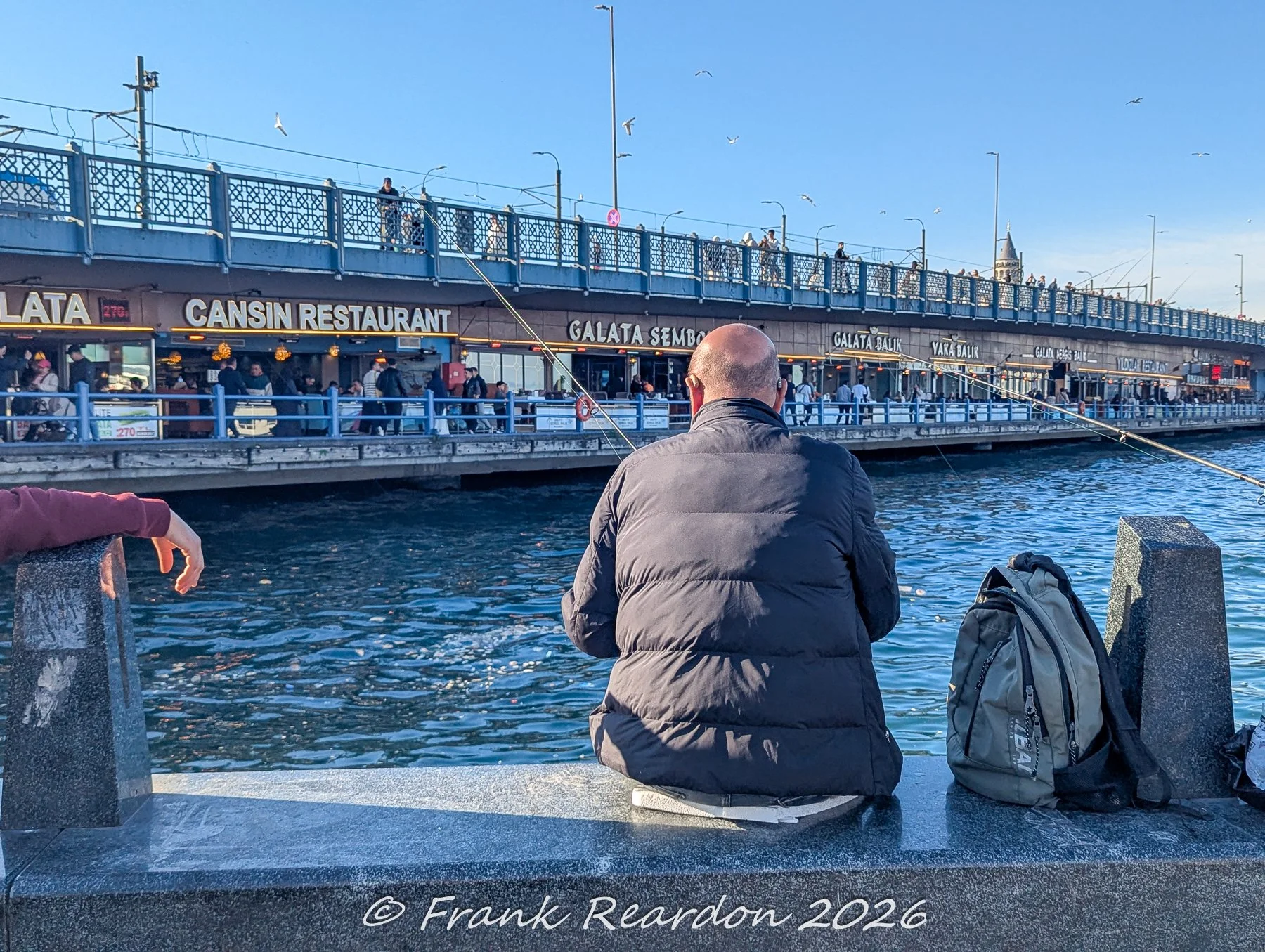 A fisherman sitting on the stone quay with his back turned, casting his line toward the Galata Bridge and its lower-deck restaurants like Cansin Restaurant and Galata Balık.