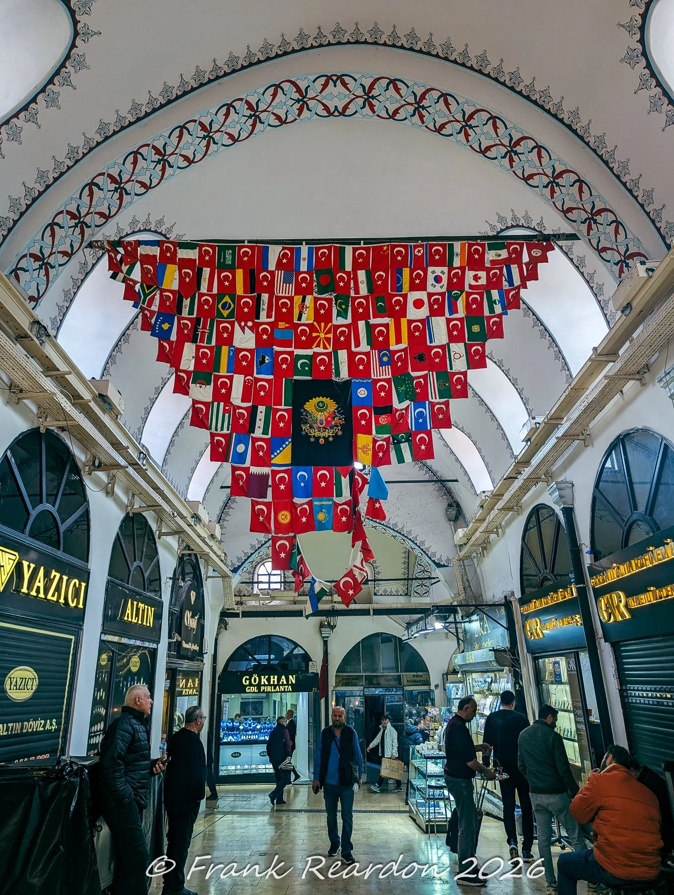 A colorful array of international flags and an Ottoman imperial crest suspended from the arched, painted ceiling of a busy shopping arcade within the Grand Bazaar.