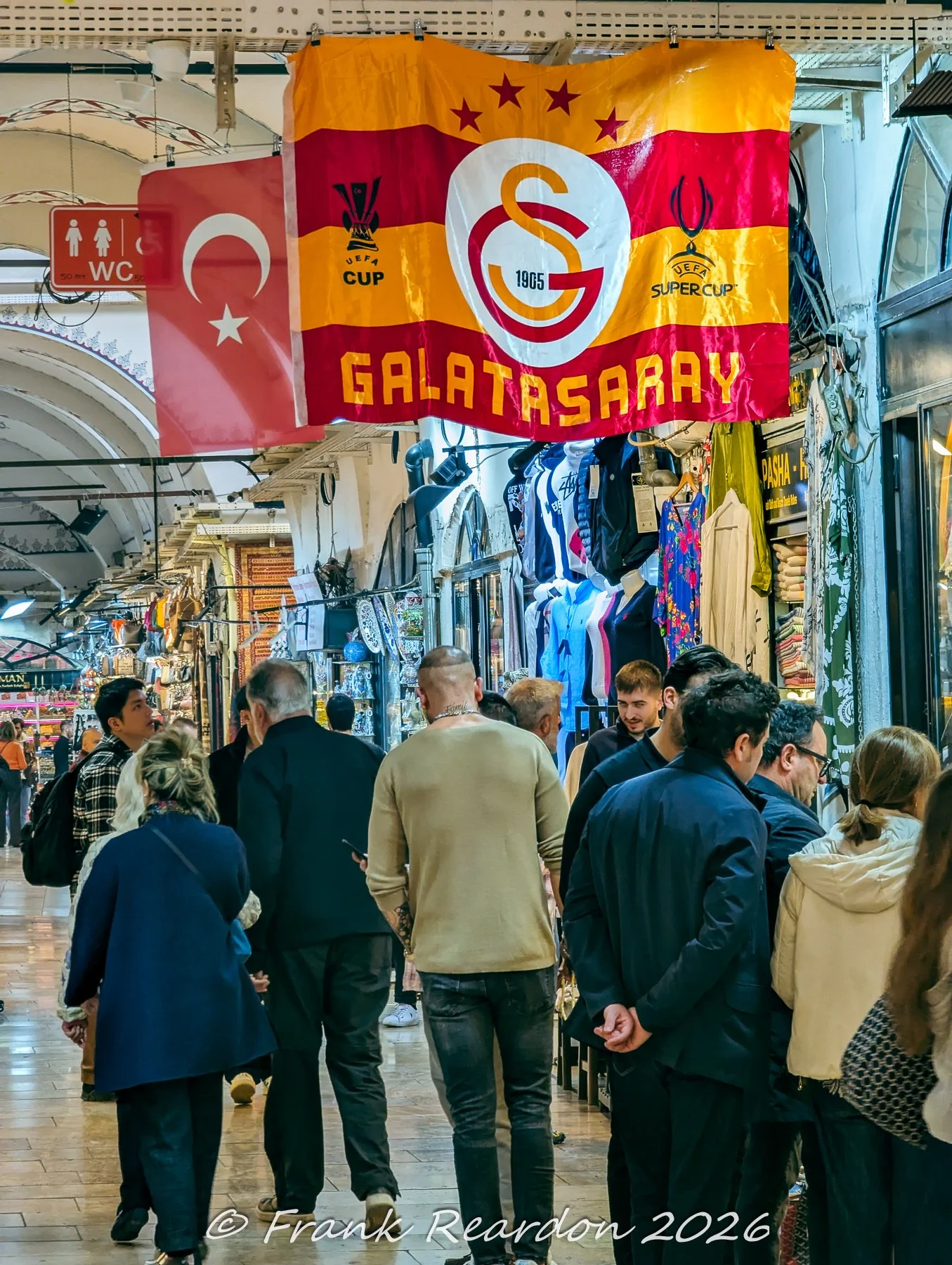 A vibrant Galatasaray sports club flag hangs proudly above a bustling corridor filled with shoppers and apparel stalls inside the Grand Bazaar (Kapalıçarşı)