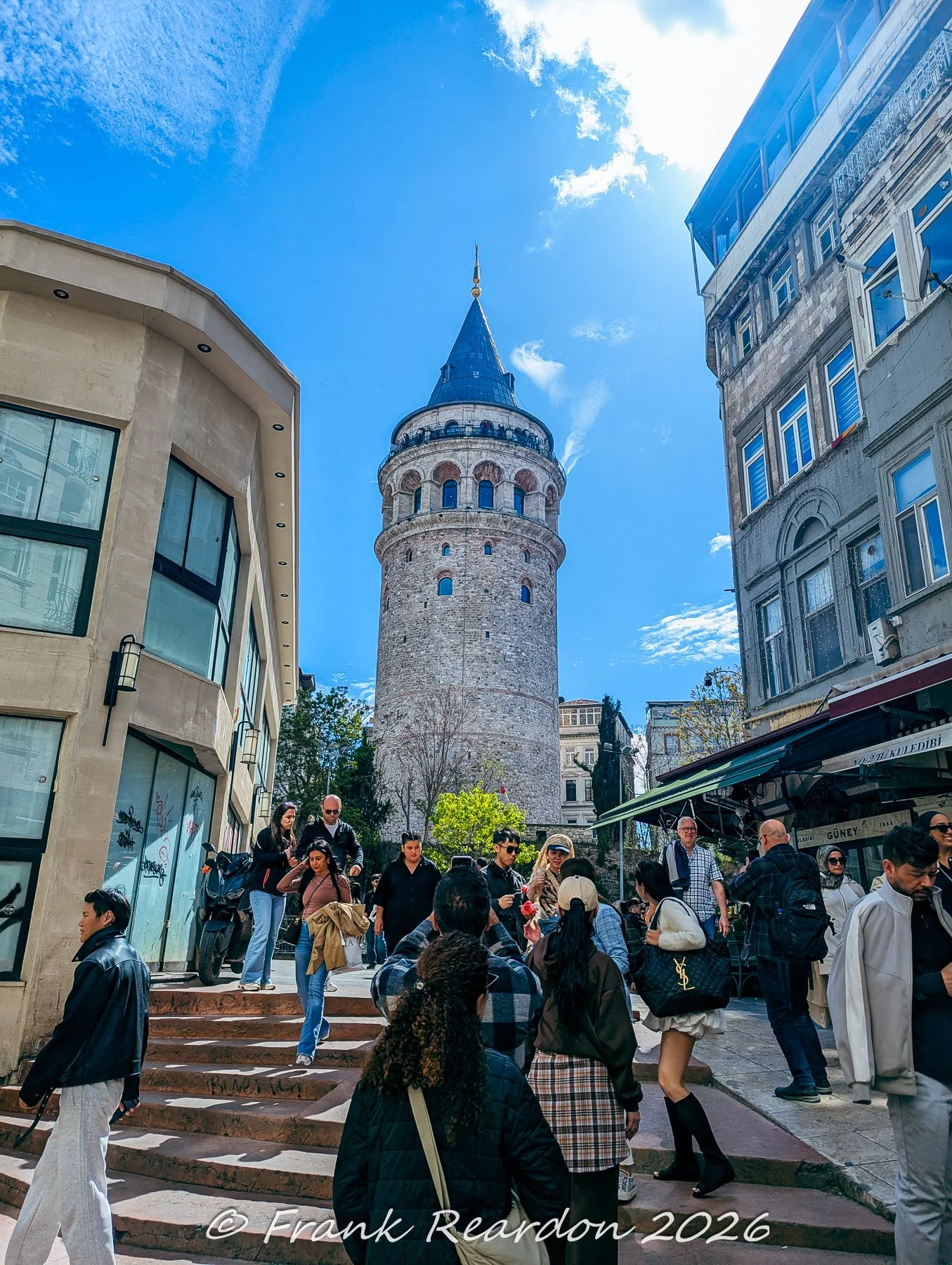 Pedestrians walking along the stone steps and bustling streets leading up to the historic Galata Tower under a bright blue sky.