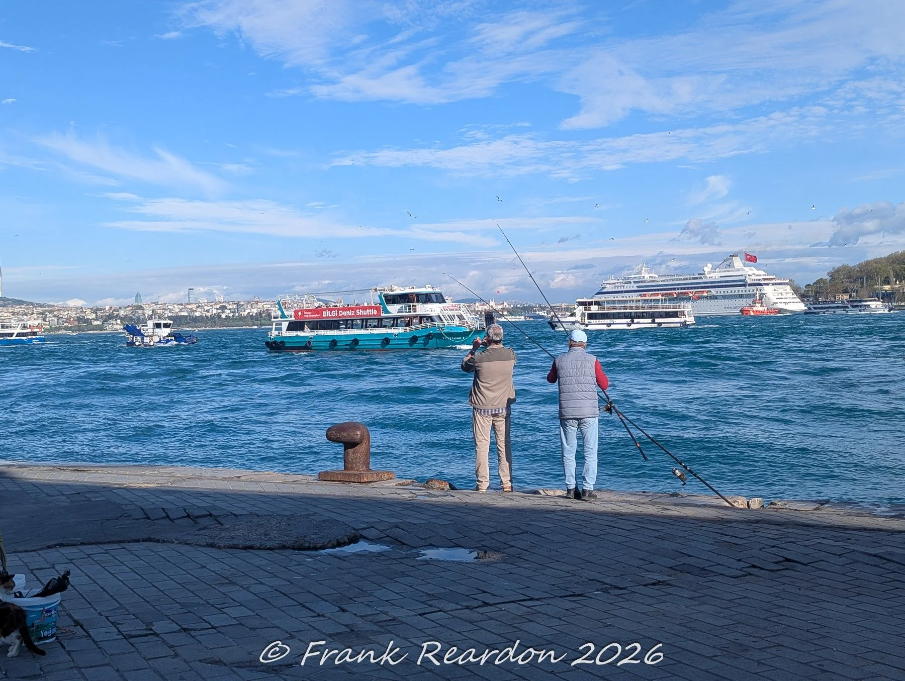 A wide daytime shot of the busy Bosphorus strait filled with various passenger ferries and shuttle boats, with fishermen standing along the paved quay.
