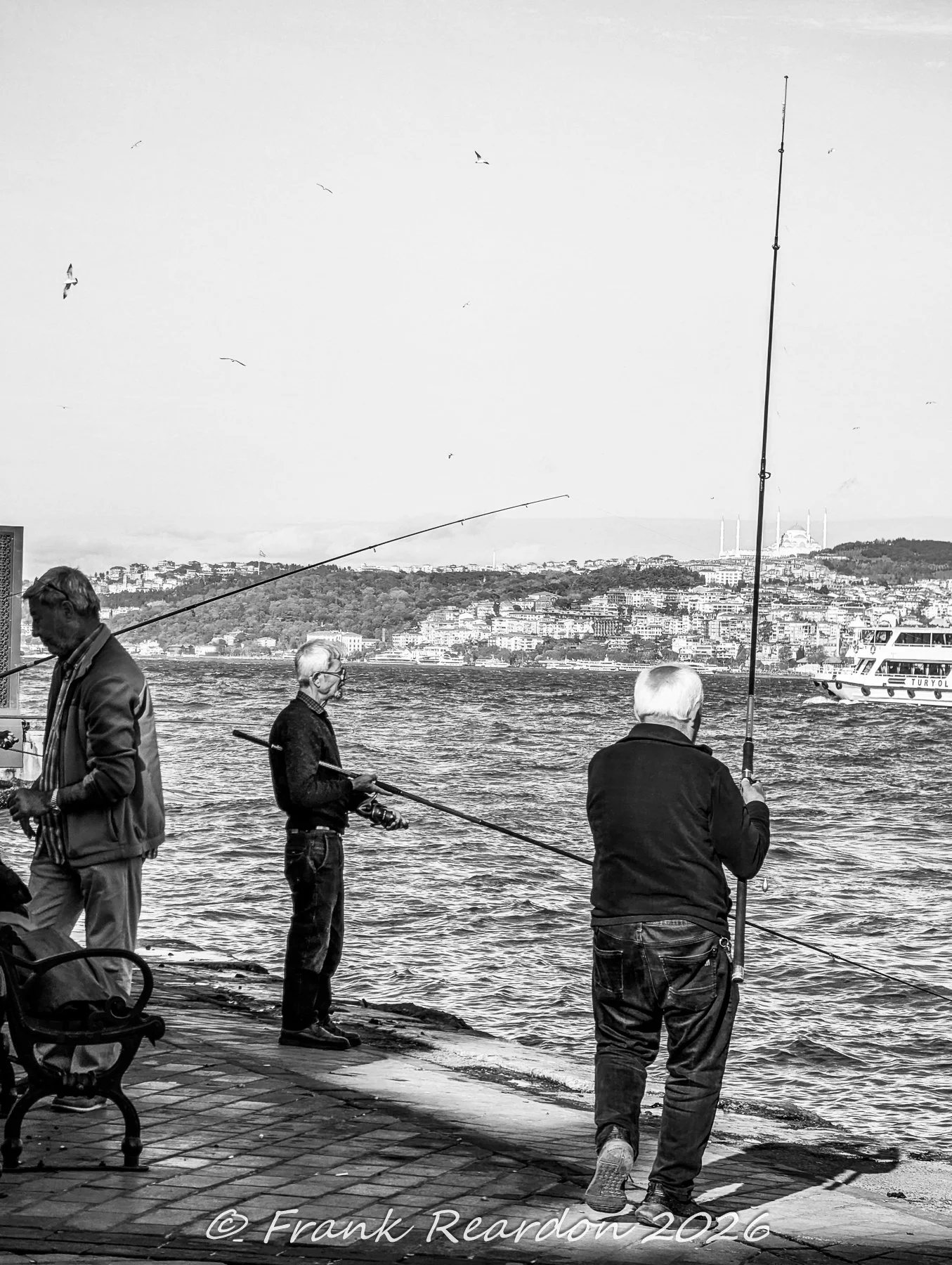 A black-and-white composition capturing local fishermen casting their lines along the waterfront with distant hills and minarets on the horizon.