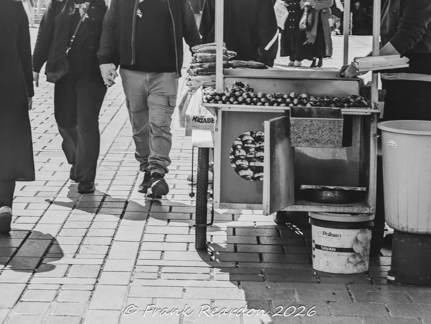 A black-and-white street scene showing pedestrians walking along a paved urban sidewalk past a traditional vendor cart.