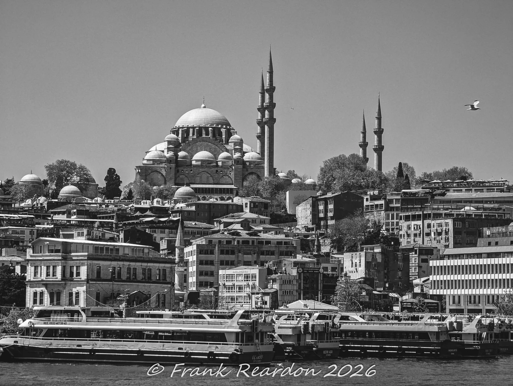 A classic monochrome architectural photograph featuring the magnificent domes and towering minarets of the historic Süleymaniye Mosque overlooking the waterfront ferry docks.