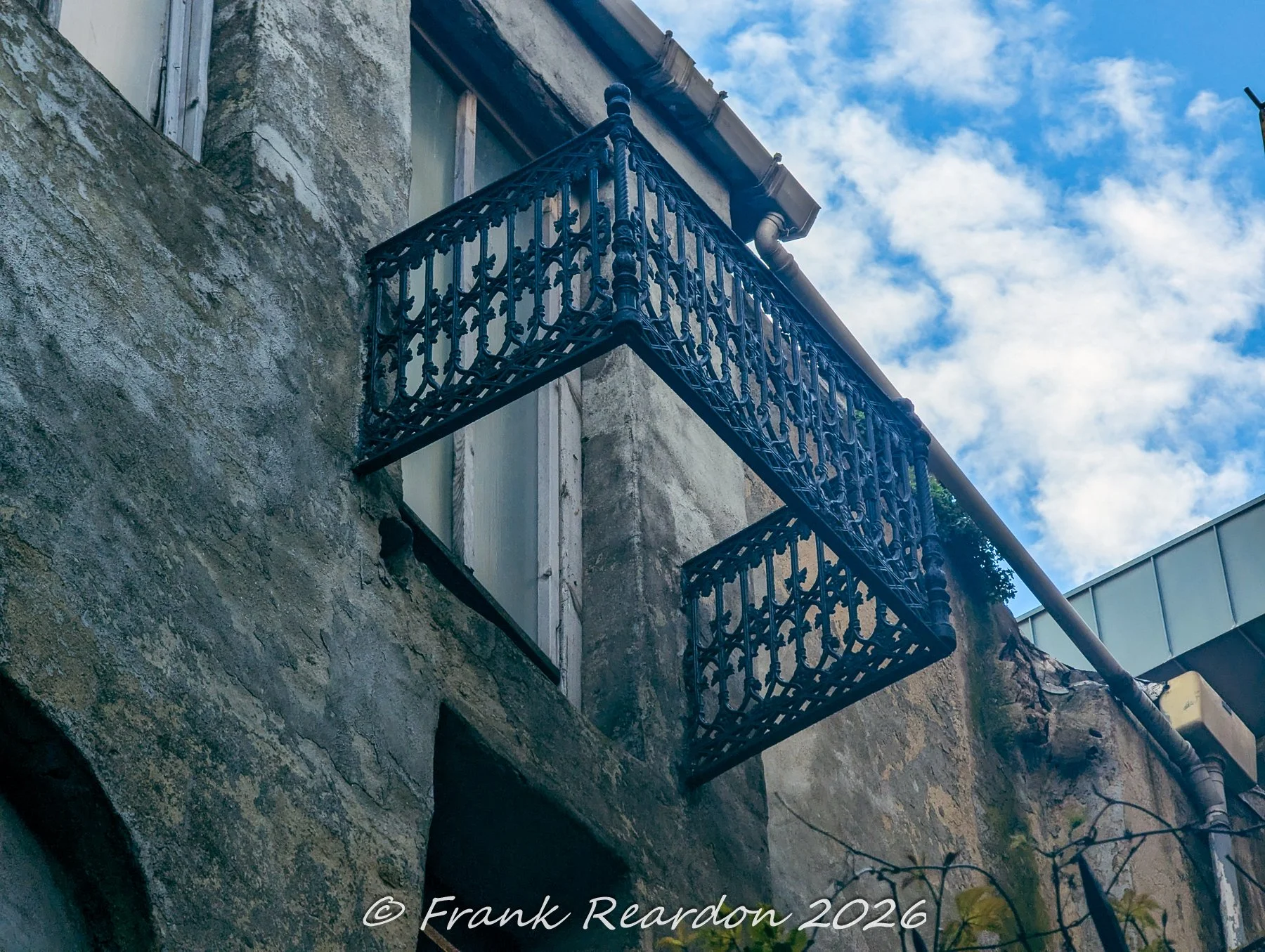 A close-up view of an elaborate, dark ornamental iron railing on a weathered, aged building exterior.