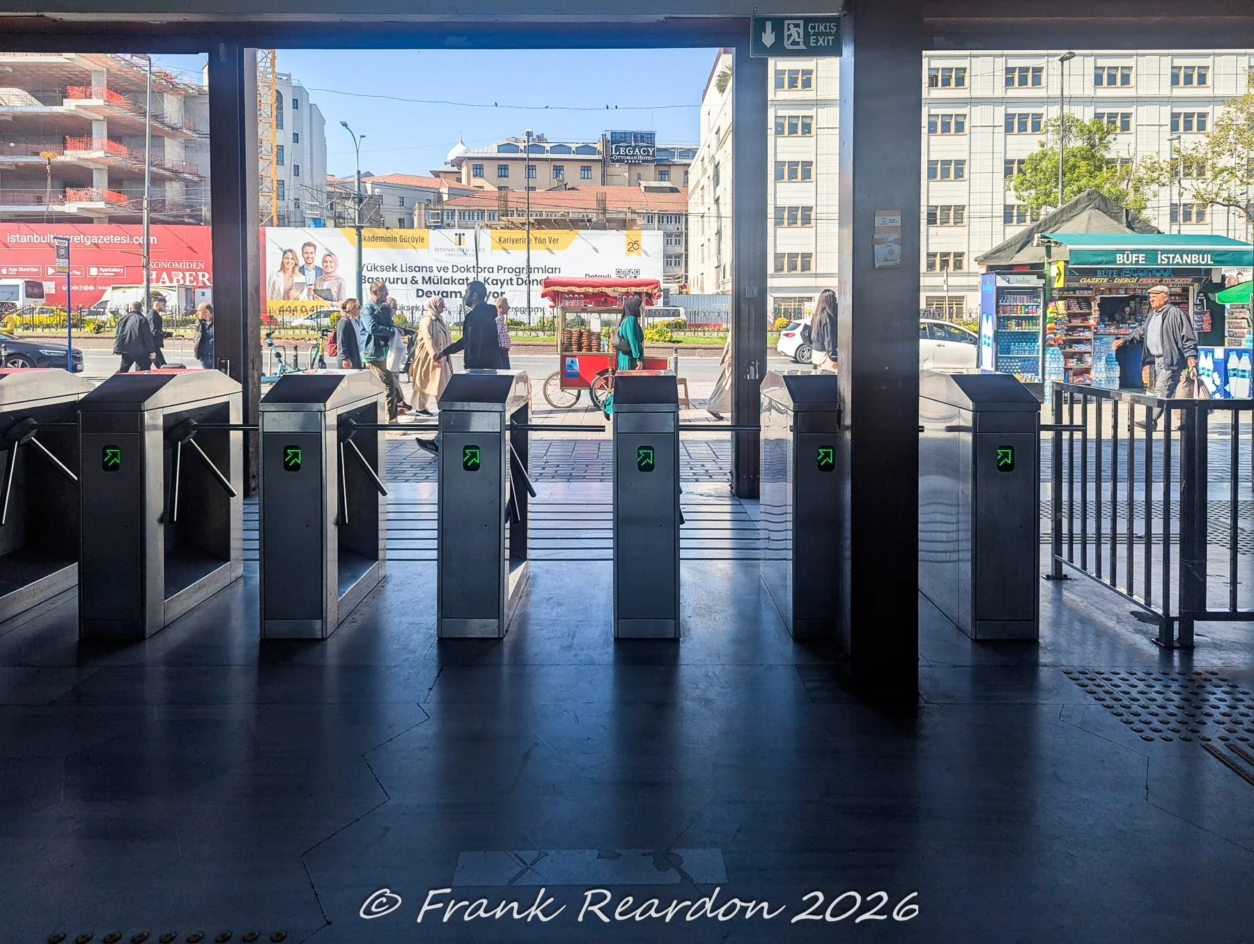 Looking out through the metal transit turnstiles and exit gates of Sirkeci Station toward the bustling street, outdoor kiosks, and urban storefronts.