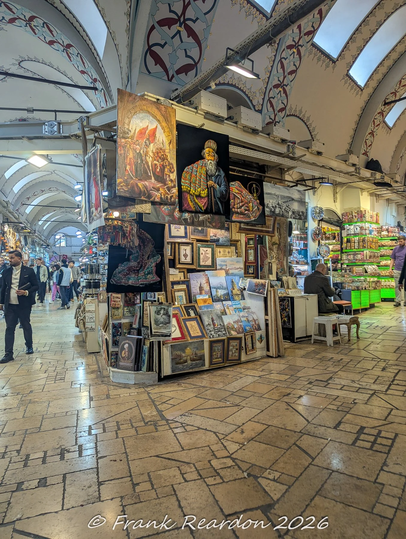 Framed paintings, portraits, and traditional artwork displayed on a central island kiosk beneath the decorated vaulted ceilings of the Grand Bazaar.
