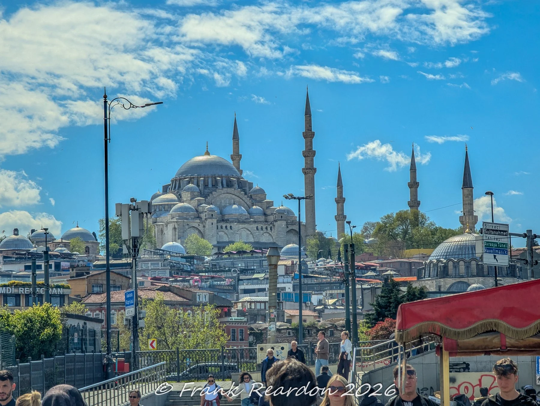 A panoramic view showcasing the grand domes and towering minarets of the historic Süleymaniye Mosque overlooking the urban landscape and street-level activity below.