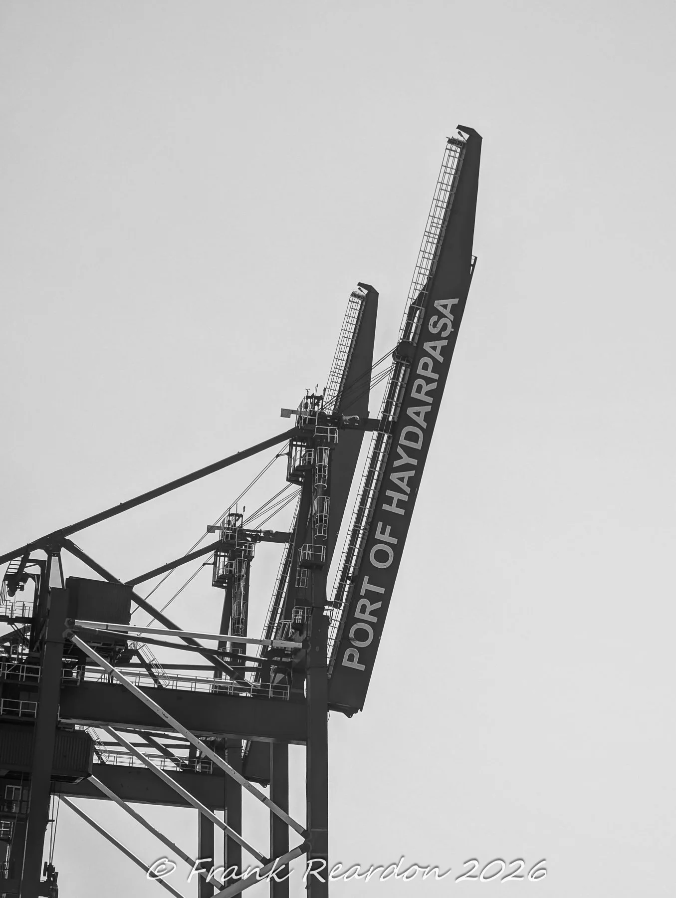 A high-contrast, black-and-white low-angle shot focusing on the massive steel lattice framework of a container crane clearly marked with "PORT OF HAYDARPAŞA".