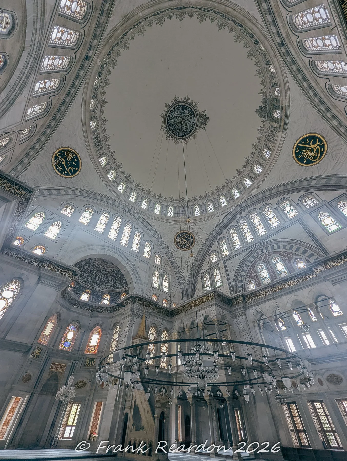 A wide-angle interior view showing the large circular chandelier hanging beneath the massive dome and arched windows of the Nuruosmaniye Mosque.