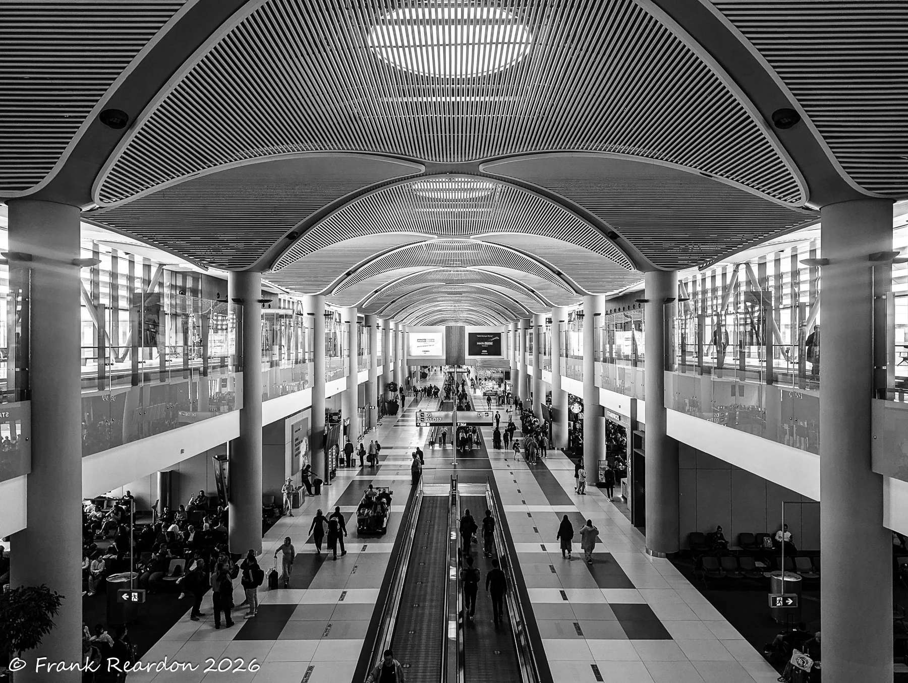 A dramatic monochrome wide-angle perspective of the bustling multi-level terminal concourse, showcasing striking vaulted ceilings, walkways, and travelers on the move.