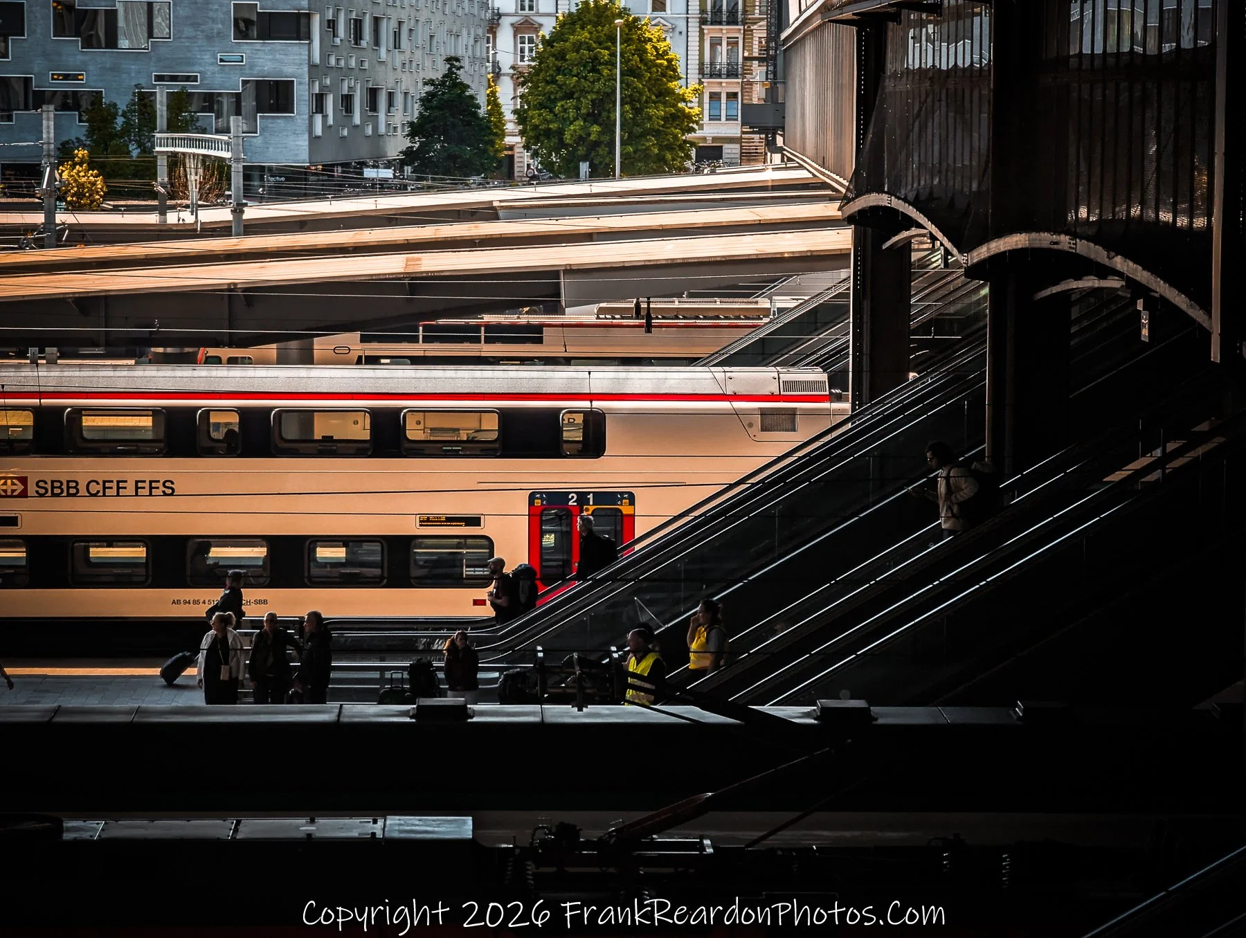 "Station Commute" A shadowed view of commuters using an escalator, contrasted with the bright, sunlit train cars waiting on the adjacent track.