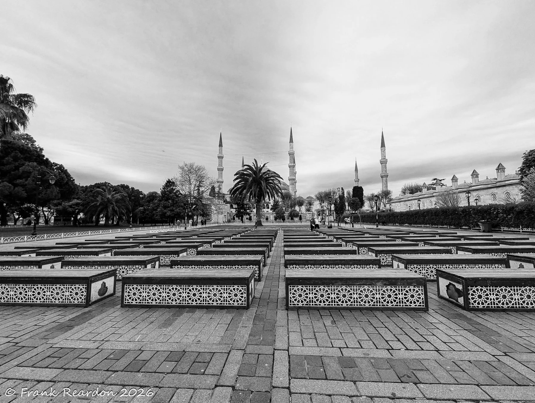 A black and white view across an orderly courtyard filled with patterned benches toward majestic mosque minarets under a cloudy sky.
