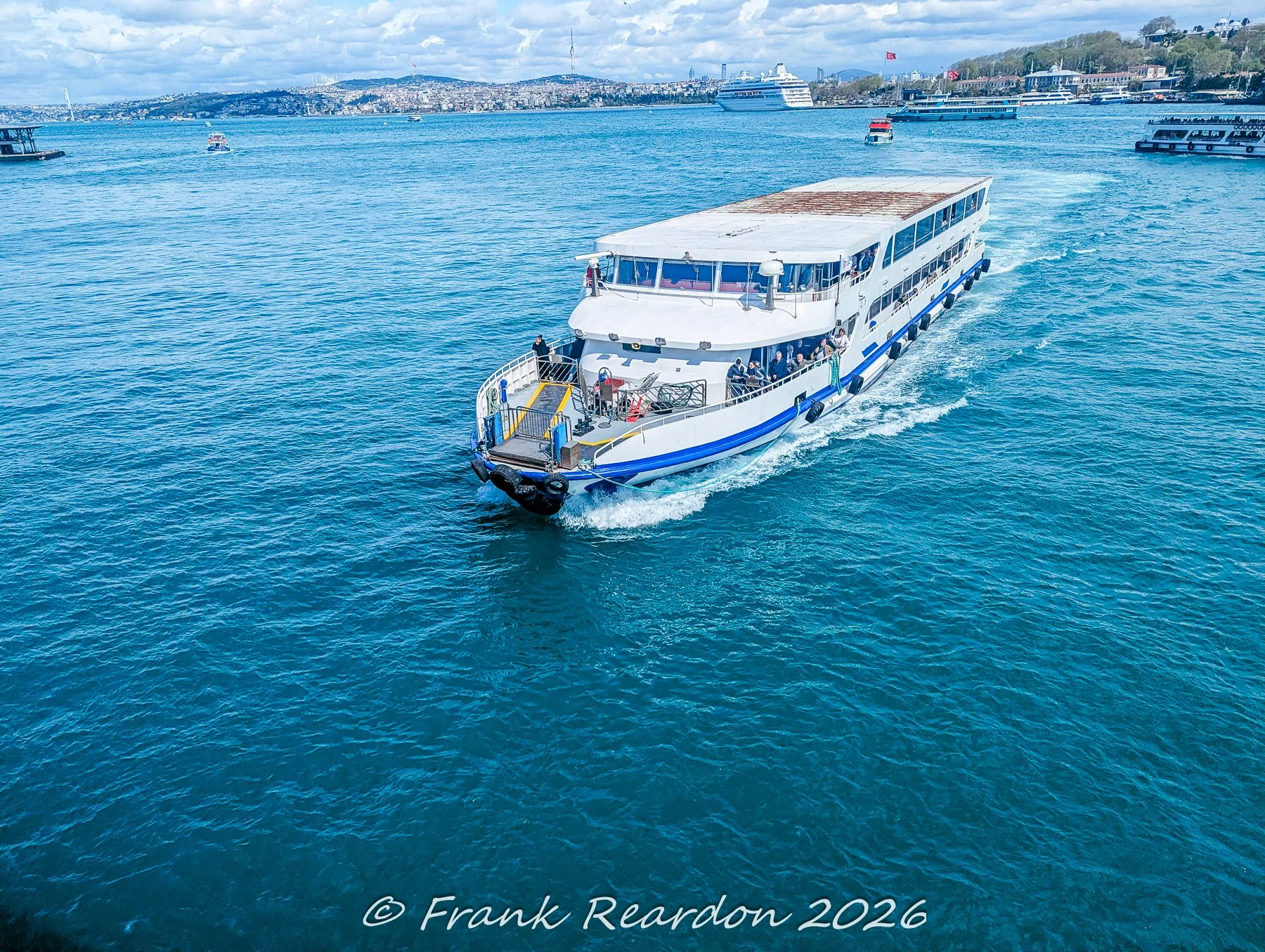 A passenger ferry navigating the bright blue waters of the Bosphorus strait in Istanbul, with distant hills and other vessels visible across the horizon.