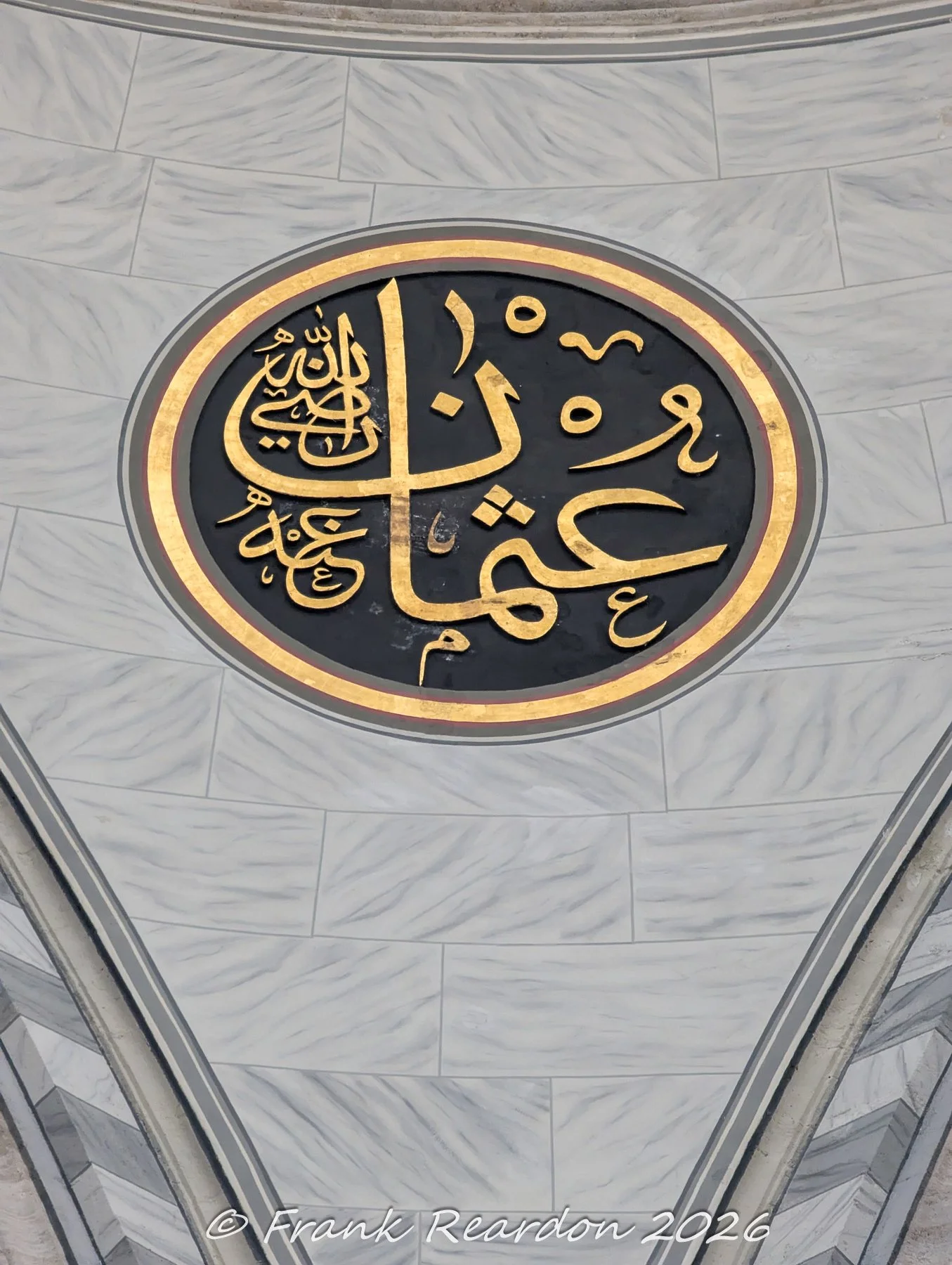 detailed close-up of a large black-and-gold calligraphic medallion mounted on the polished marble wall inside the Nuruosmaniye Mosque. "displays the name Uthman (عثمان), referring to Uthman ibn Affan, the third Caliph of Islam and a close companion o