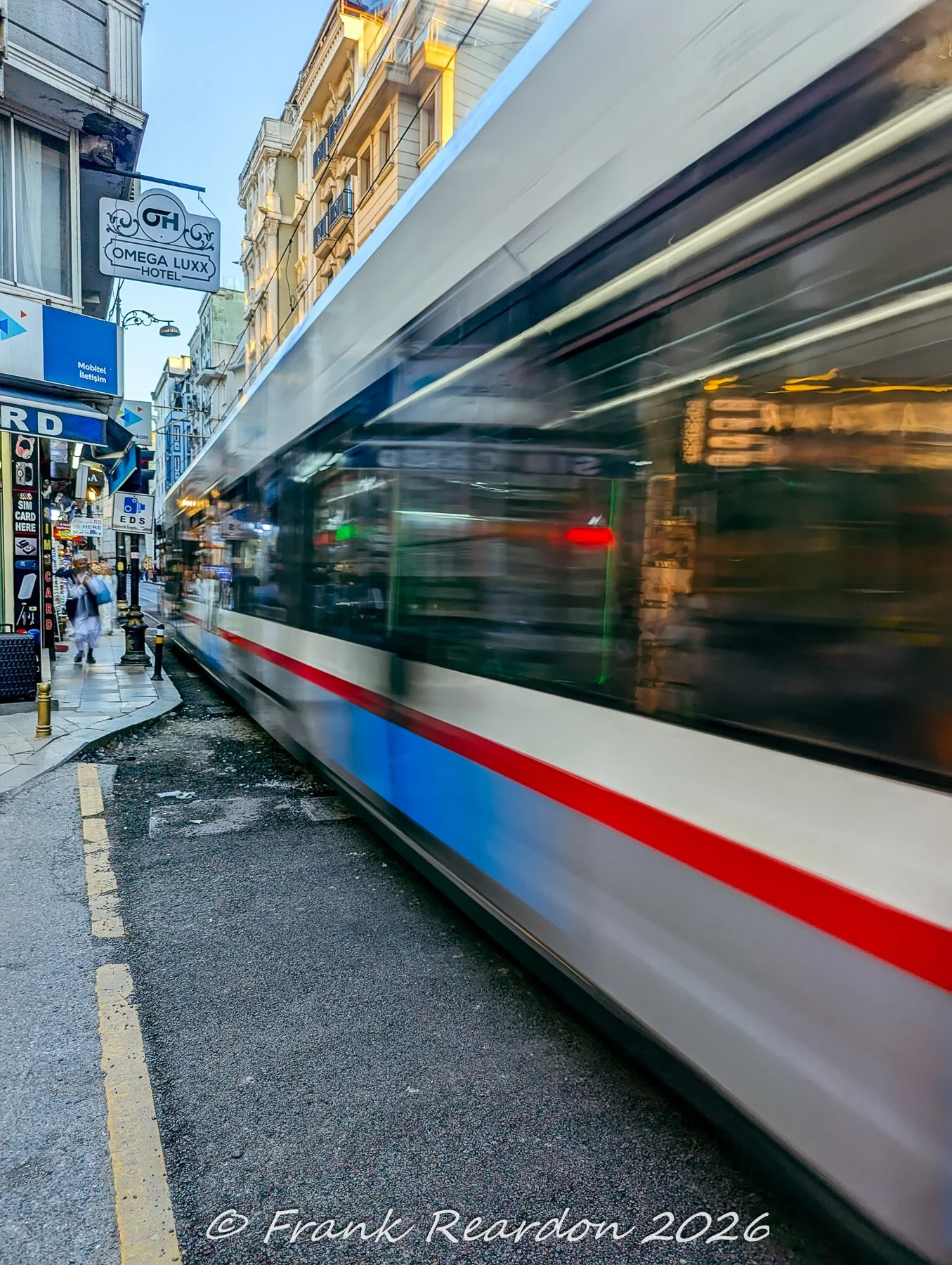 A long-exposure action shot capturing a modern urban tram rushing past storefronts, along the narrow historic streets.