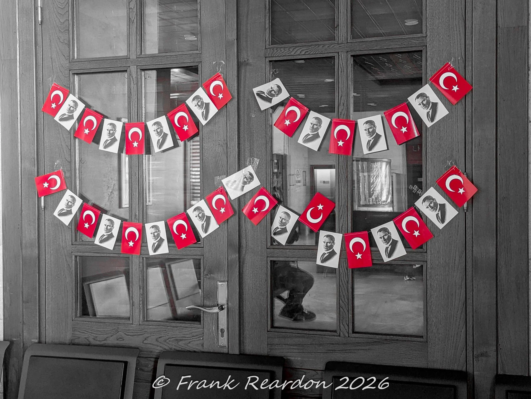 A black-and-white close-up of wooden glass doors adorned with festive strings of Turkish flags and portraits of Mustafa Kemal Atatürk inside Sirkeci Station.