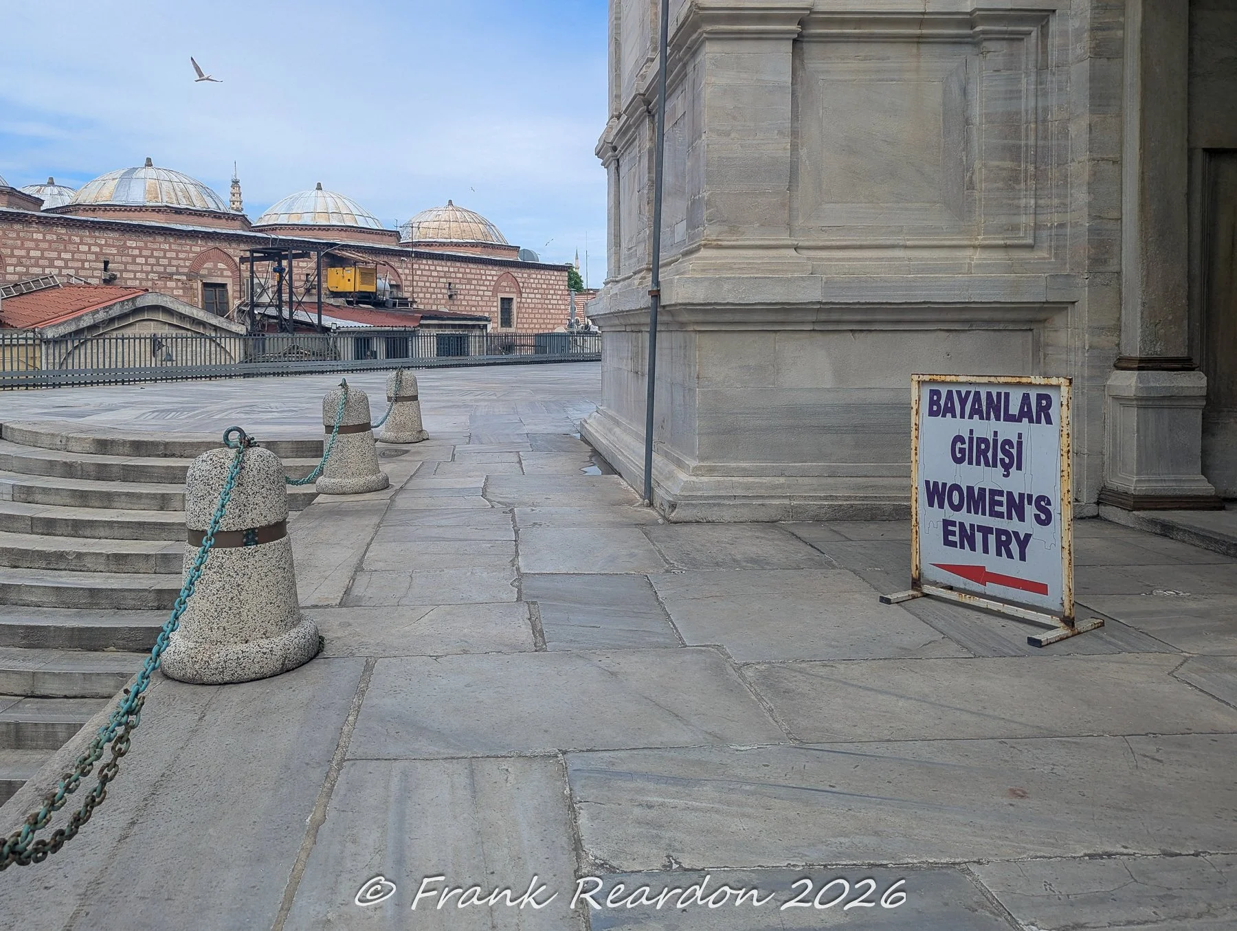 A directional sign indicating the "Women's Entry" (Bayanlar Girişi) on the stone-paved terrace overlooking the domes and rooftops surrounding the Nuruosmaniye Mosque.