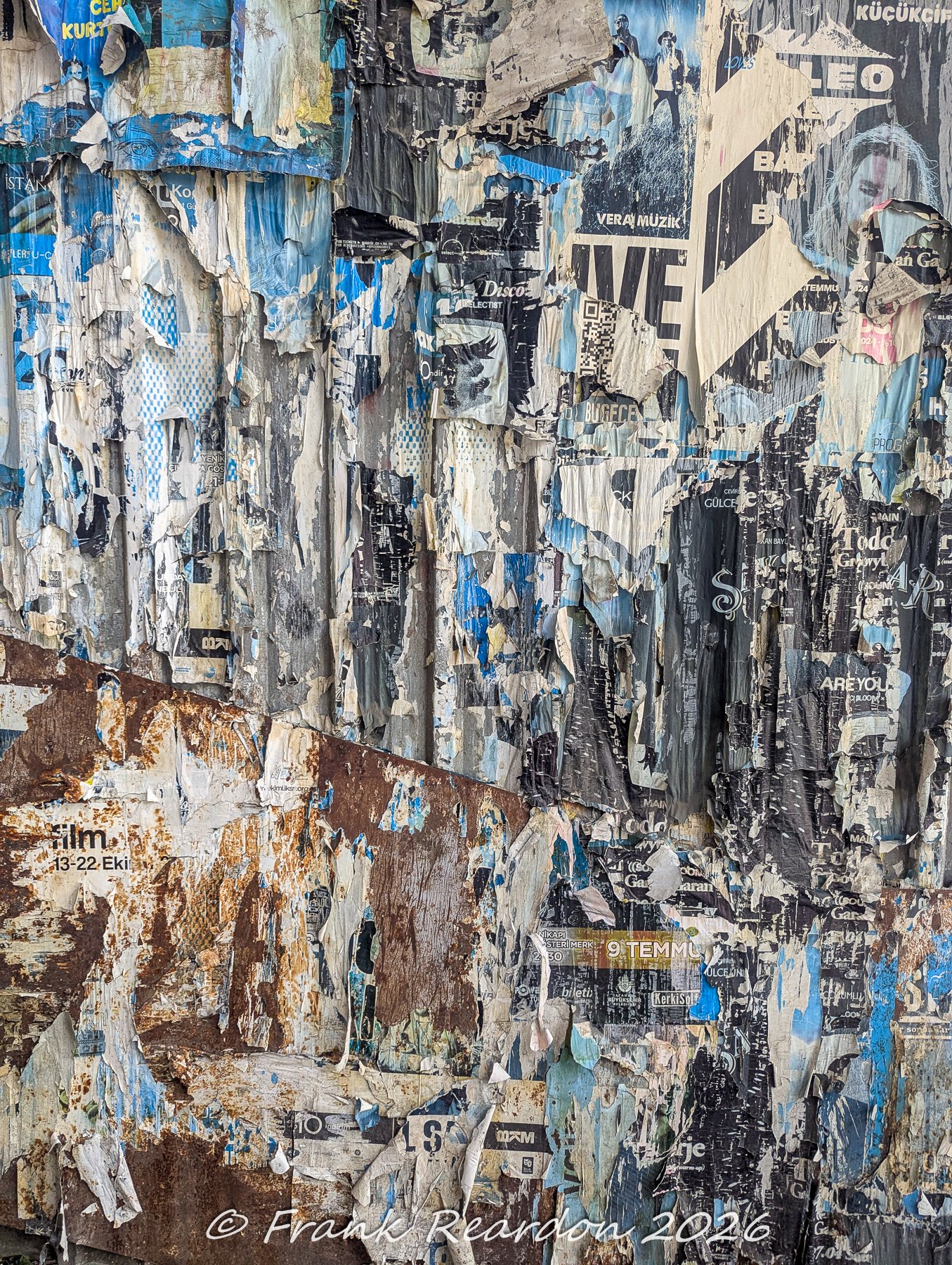 A close-up texture shot of a weathered city wall covered in layers of torn concert and event posters peeling over rusted metal.