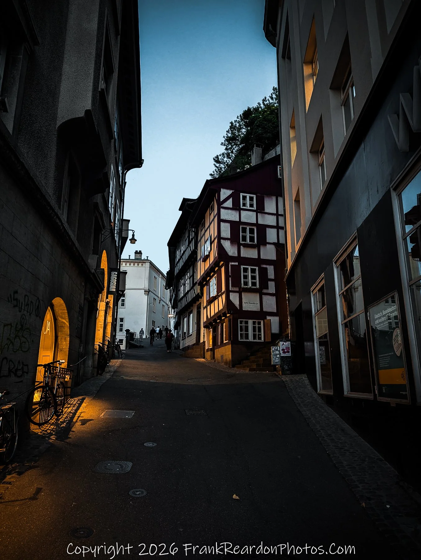 "Basel Golden Hour" A narrow, picturesque street scene at golden hour, where warm artificial light spills from a doorway onto the pavement, illuminating a charming timber-framed building.