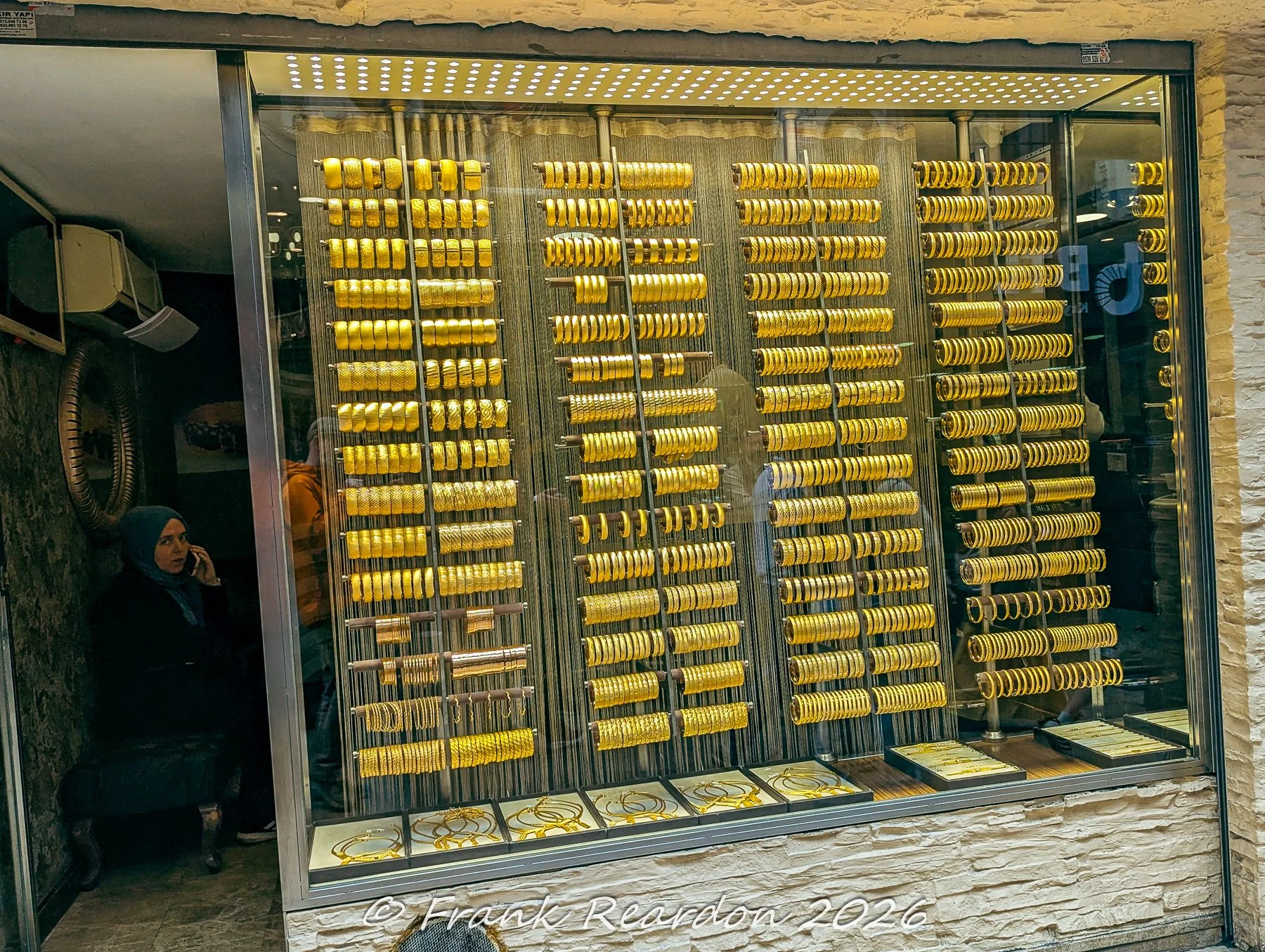 A close-up view through a secure glass window showcasing vertical rows of traditional gold bangles and wedding jewelry inside a shop near the Grand Bazaar.