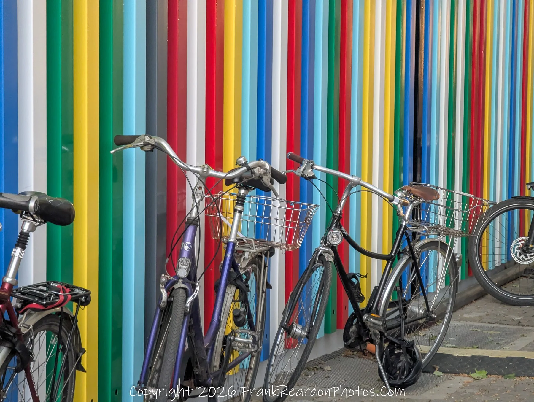 "Colorful Wall and Bicycles" Two bicycles parked against a vibrant, multi-colored wall composed of vertical panels in shades of blue, green, yellow, red, and white.