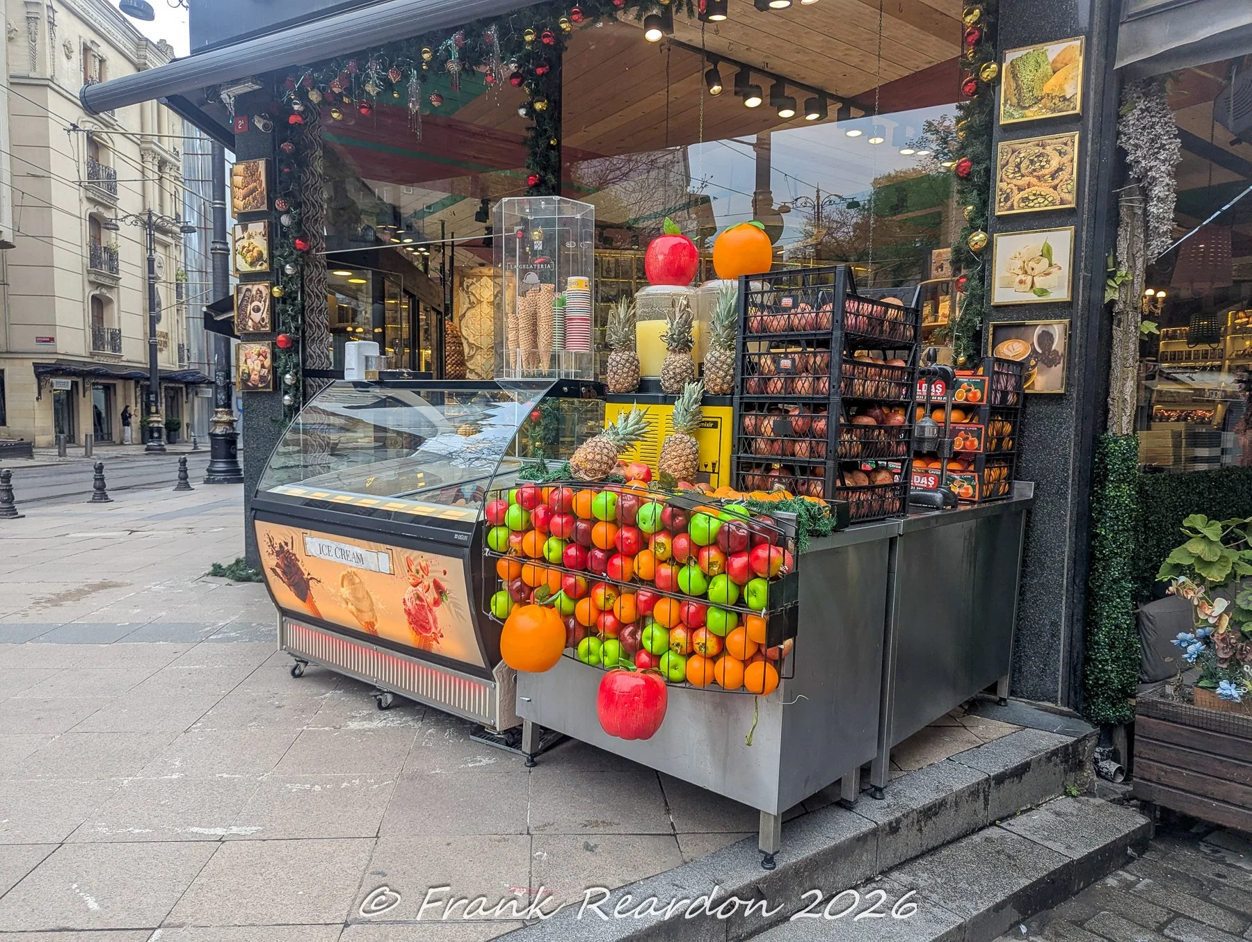 A colorful street-side vendor stand displaying fresh fruit, juice dispensers, and ice cream advertisements on a cobblestone street in Istanbul.