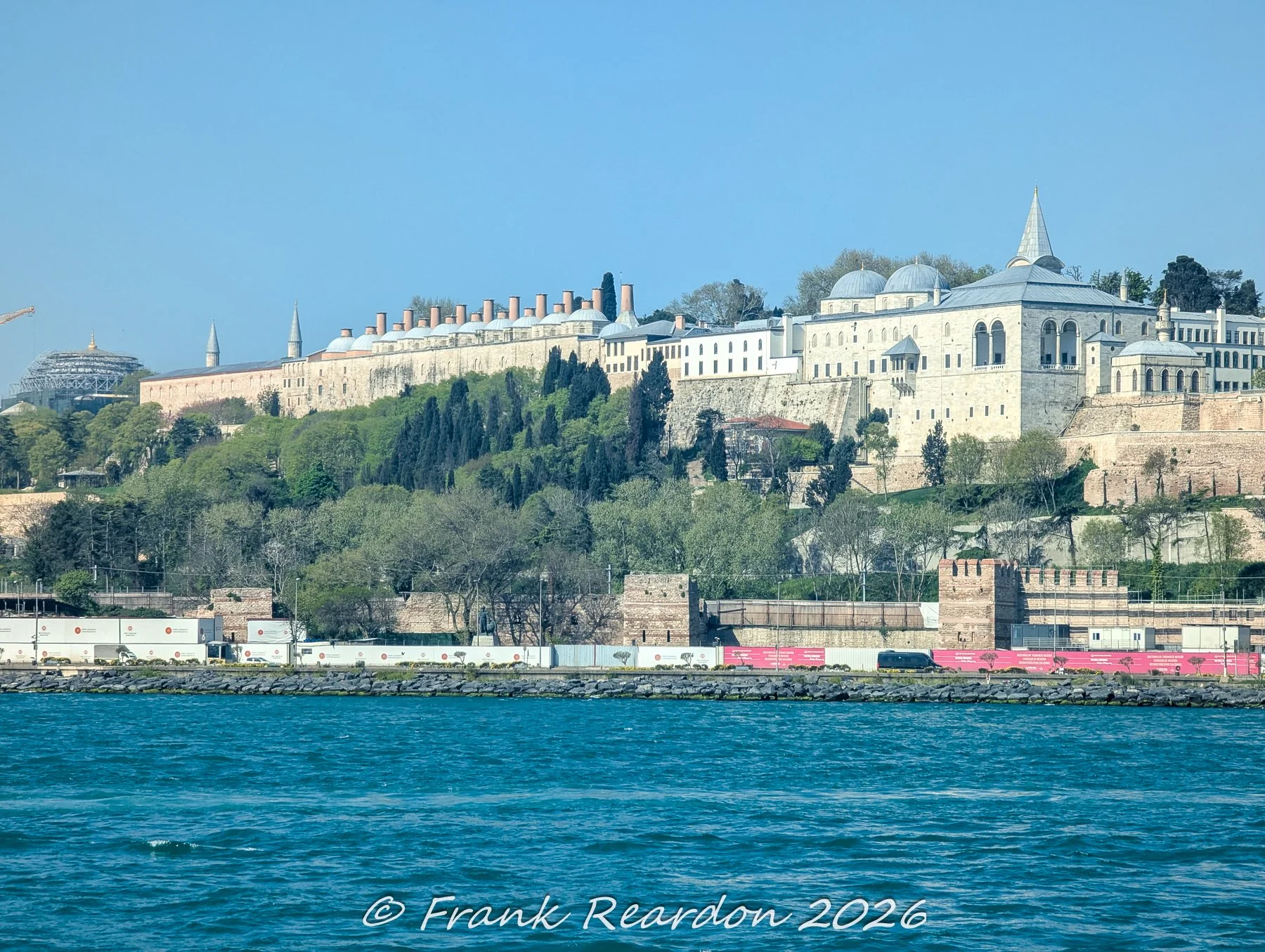 A daytime view from the water showing the majestic architecture and historic walls of Topkapi Palace rising above the coastal greenery along the Bosphorus shore.
