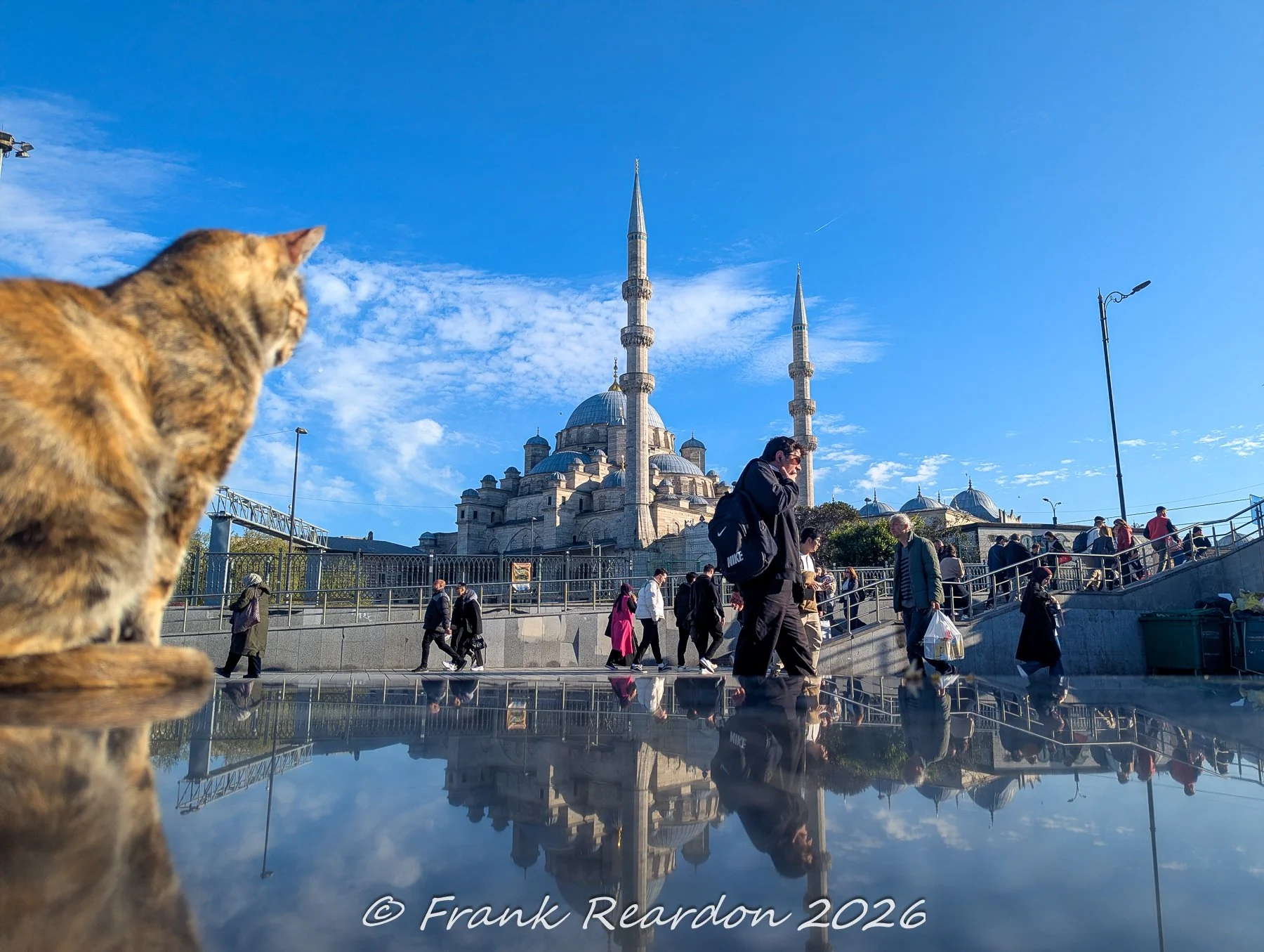 Pedestrians walking across a public square near the New Mosque (Yeni Cami), with a striking mirror-like reflection of the historic complex captured on a wet or polished surface.