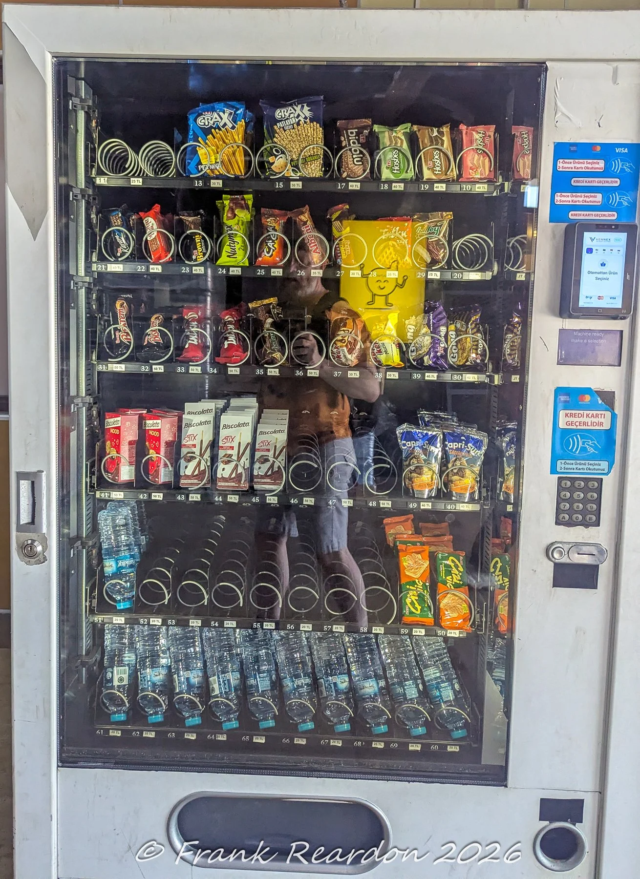 A white public vending machine stocked with various packaged snacks, chocolates, and water bottles, featuring an integrated