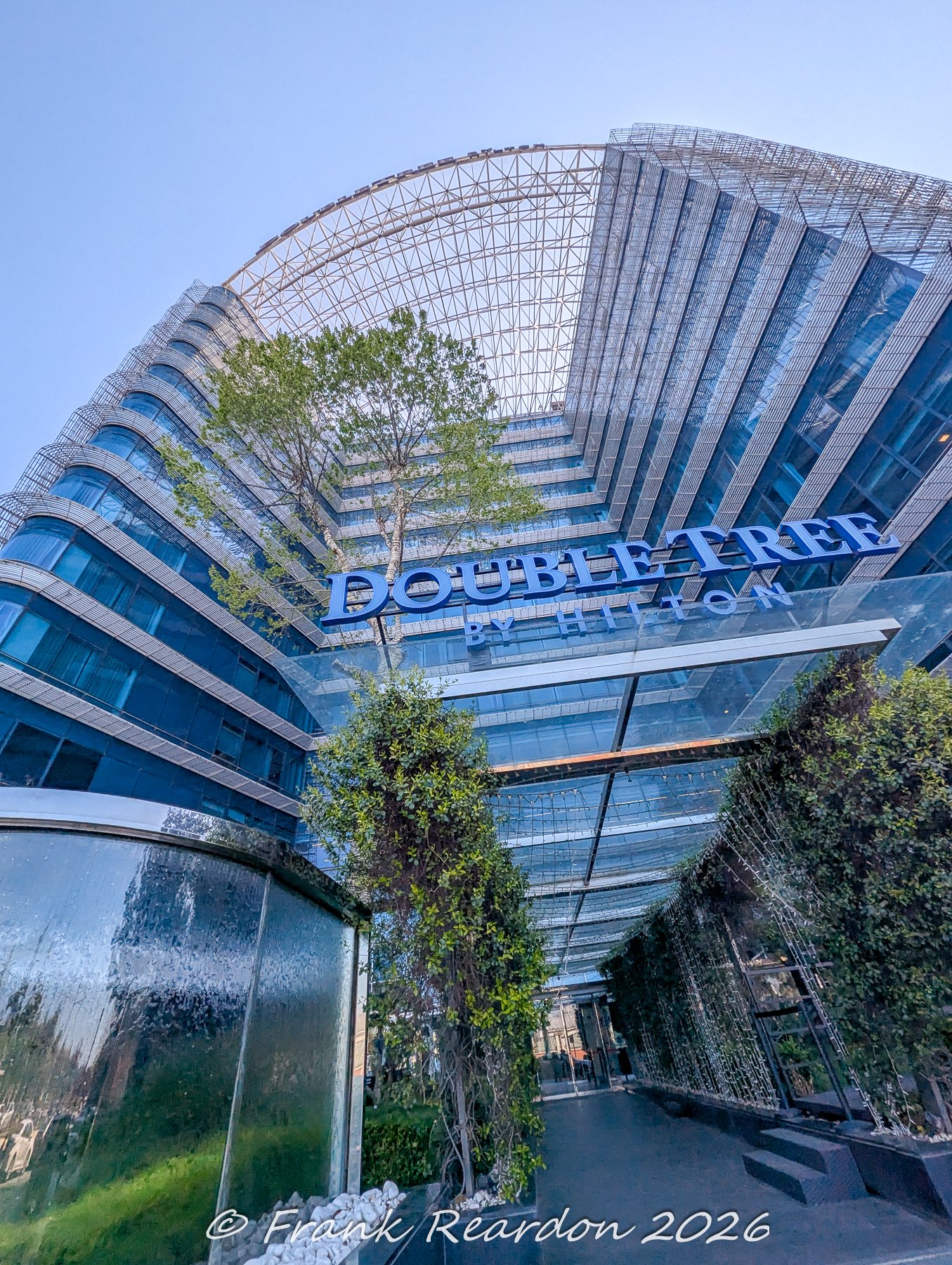 A low-angle shot looking up at the curved glass facade, arched glass canopy, and modern architectural elements of the DoubleTree by Hilton hotel.