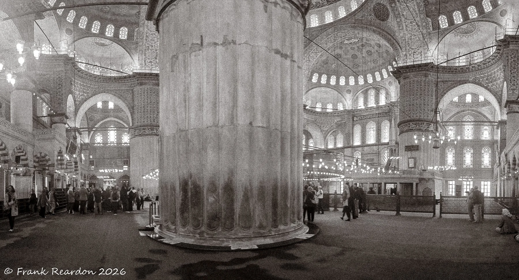 A sweeping panoramic monochrome perspective capturing the massive central pillar, vaulted arches, and visitors exploring the expansive prayer hall.