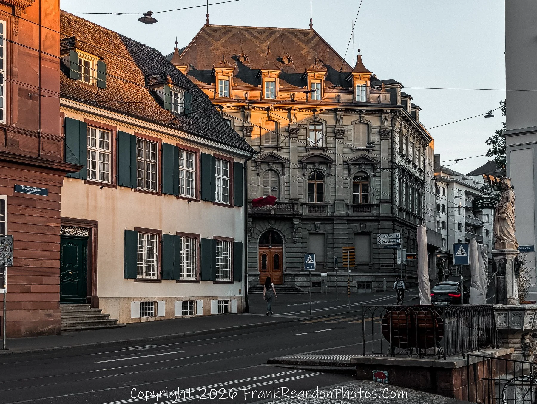 "Historic Basel Street Scene" A street-level view of an old city intersection with historic architecture, including a building bathed in warm sunset light and a stone fountain statue on the right.