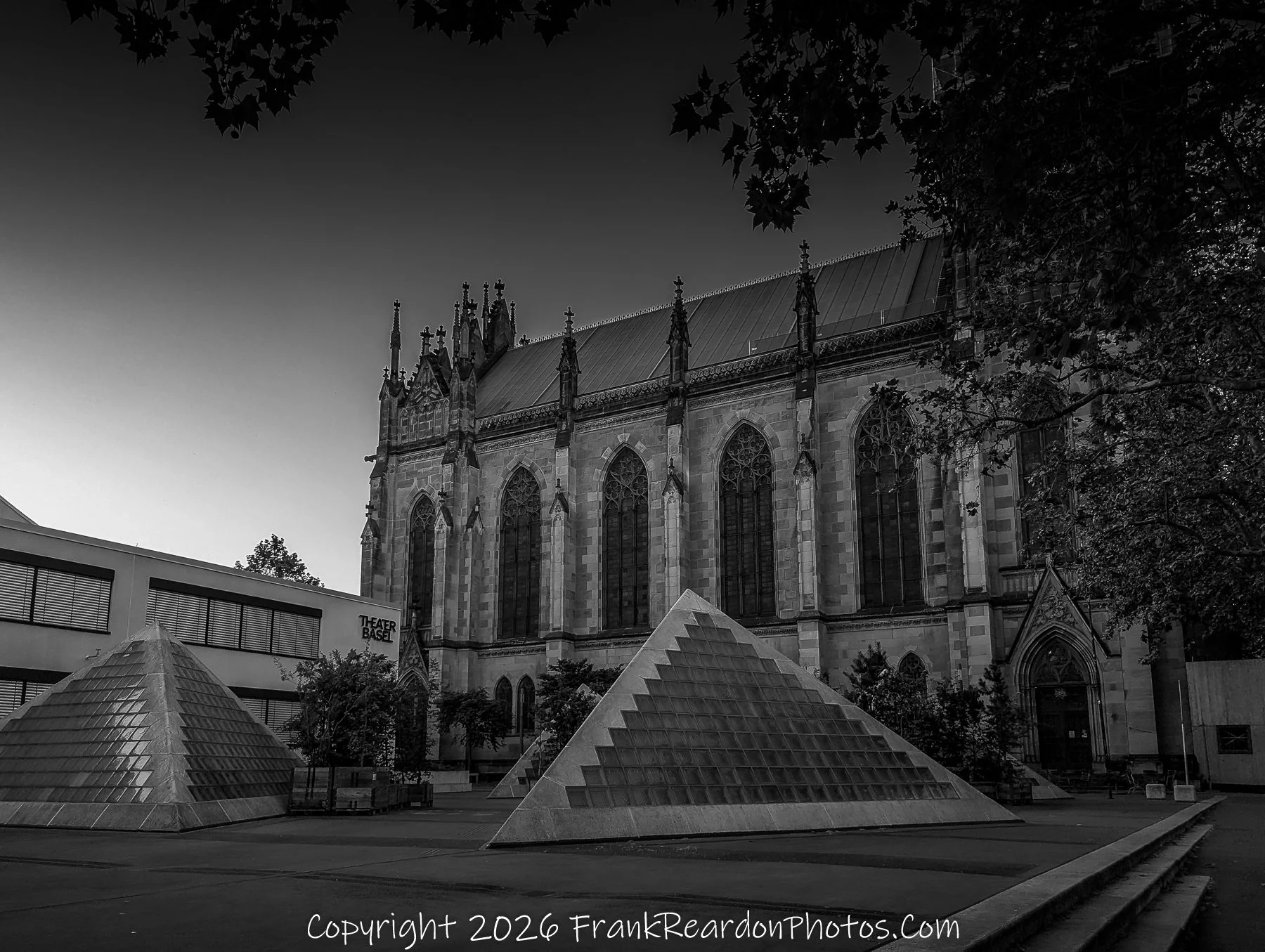 "Gothic Church and Pyramids" A wide, black-and-white shot showing an ornate, tall Gothic church building towering over the glass pyramid structures in the plaza.