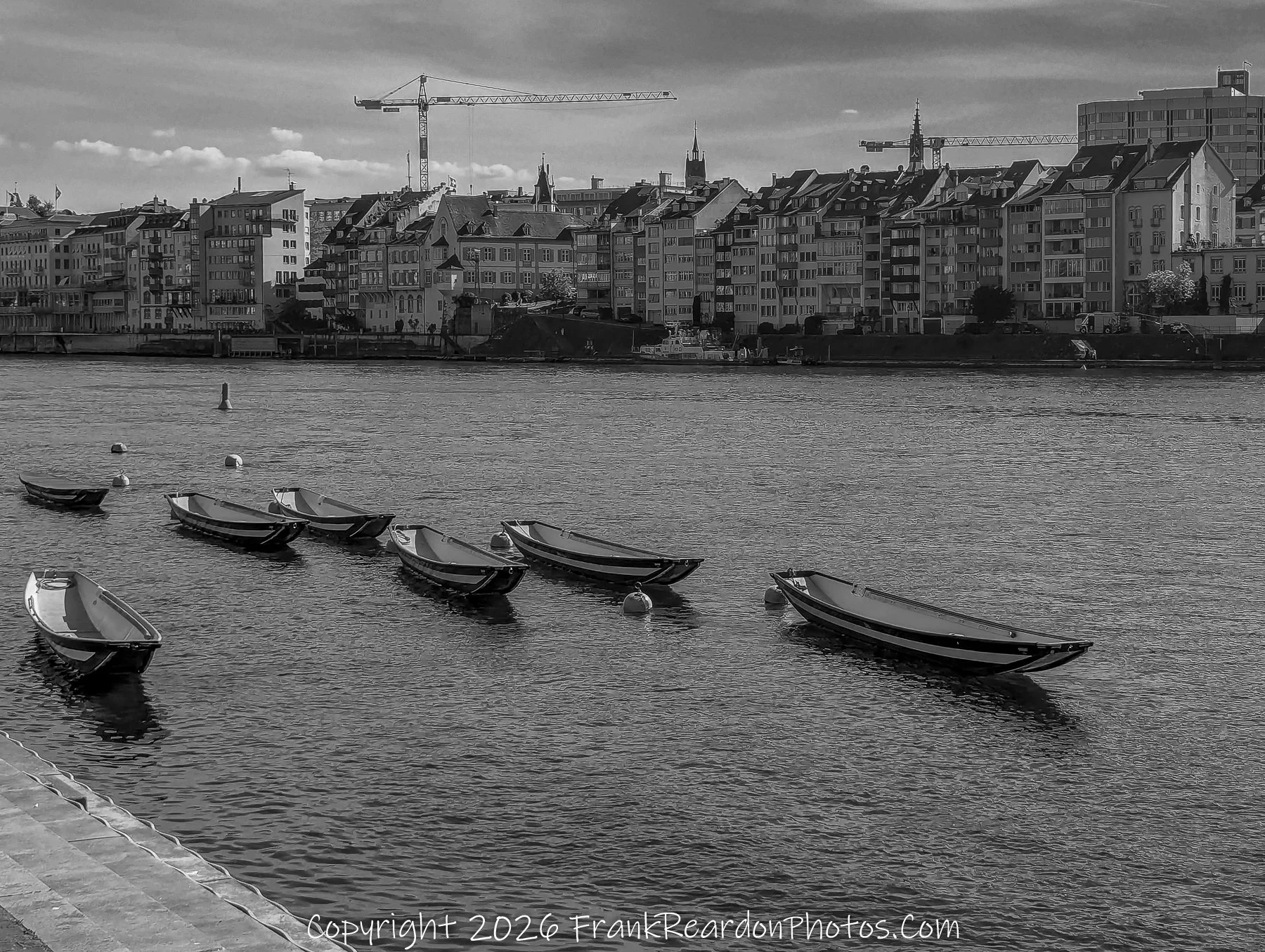 "Evening Reflections of the Basel Waterfront" A monochromatic view of small boats moored along the Rhine riverbank, with the city buildings and a construction crane in the background.