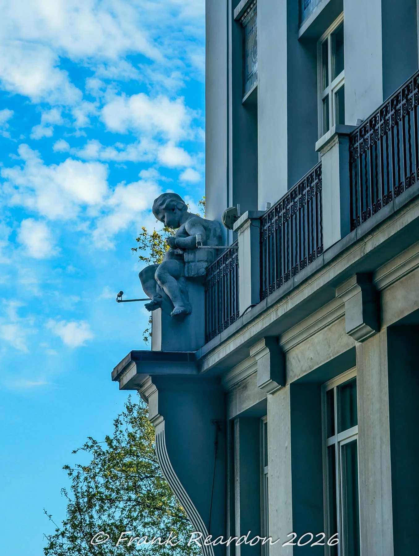 A detailed architectural shot focusing on a carved stone cherub statue perched on a corbel below a balcony railing against a blue, cloudy sky.