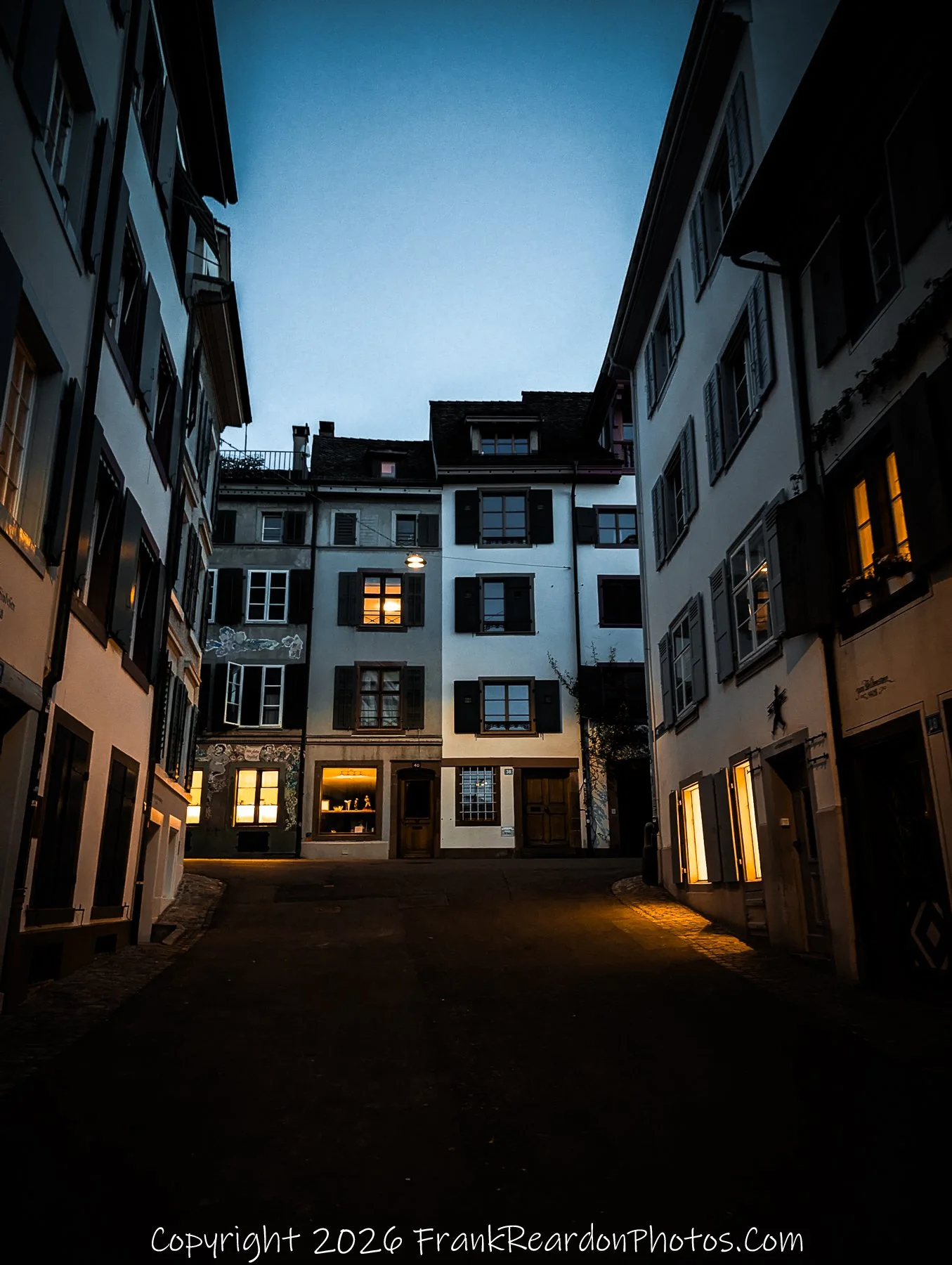 "Quiet Streets of Basel" A serene, dimly lit alleyway at night, framed by traditional multi-story buildings with glowing windows.
