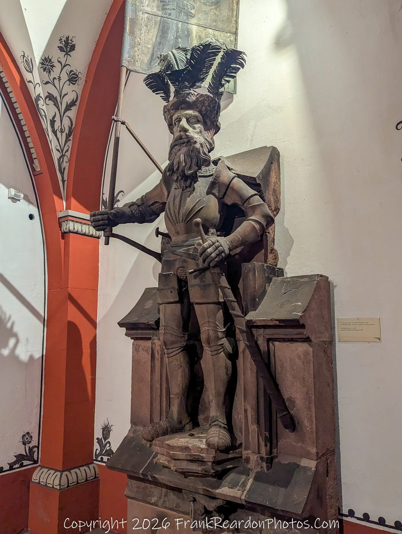 "The Knight in Stone" A detailed shot of an imposing, full-armor stone statue of a knight holding a flag, displayed in an arched, painted gallery inside the building.
