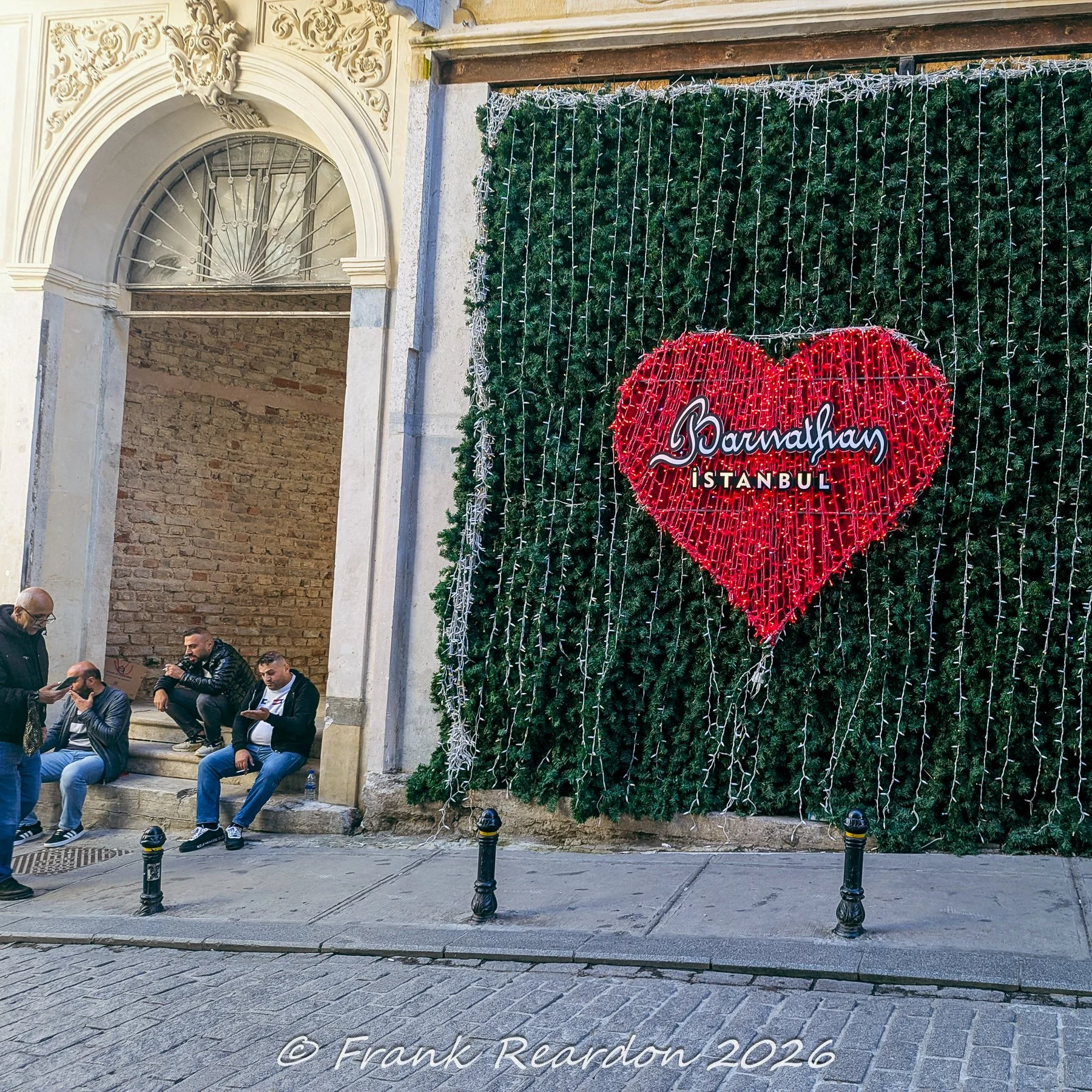 A large illuminated red heart sign featuring the text "Bermalhan Istanbul" mounted on a lush green wall outside a historic entrance, with people sitting on nearby steps.