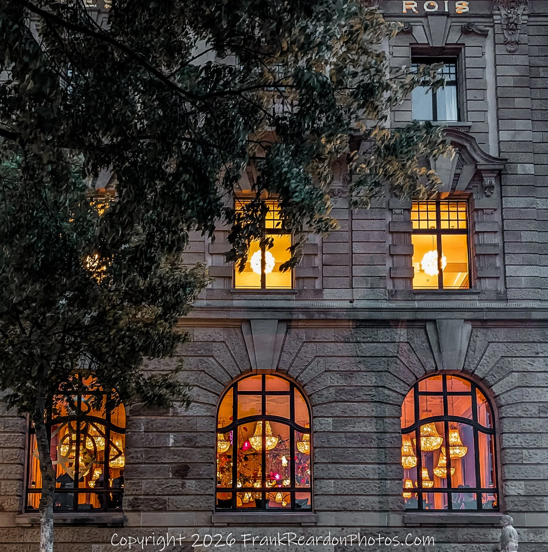 "Evening Glow at the Grand Hotel" A charming exterior view of a historic building at night, with warm, inviting light spilling from the arched windows that reveal intricate chandeliers inside.