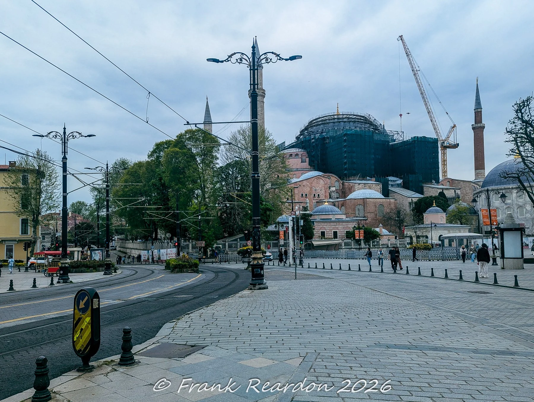 A street level view featuring the historic Hagia Sophia with its scaffolding and dome, surrounded by urban tram lines and streetlamps in Istanbul.