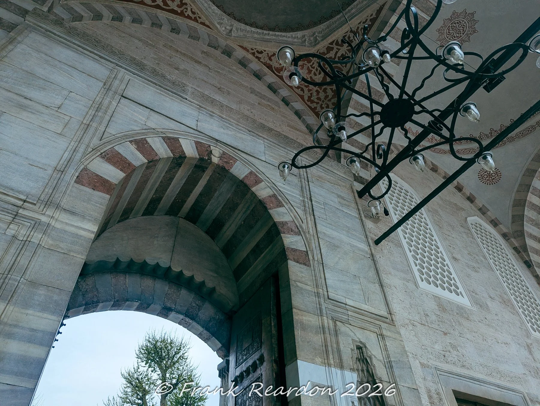 An architectural angle capturing striped stone arches, intricate tilework, and a hanging chandelier at the entrance of the Sultan Ahmed Mosque (Blue Mosque)