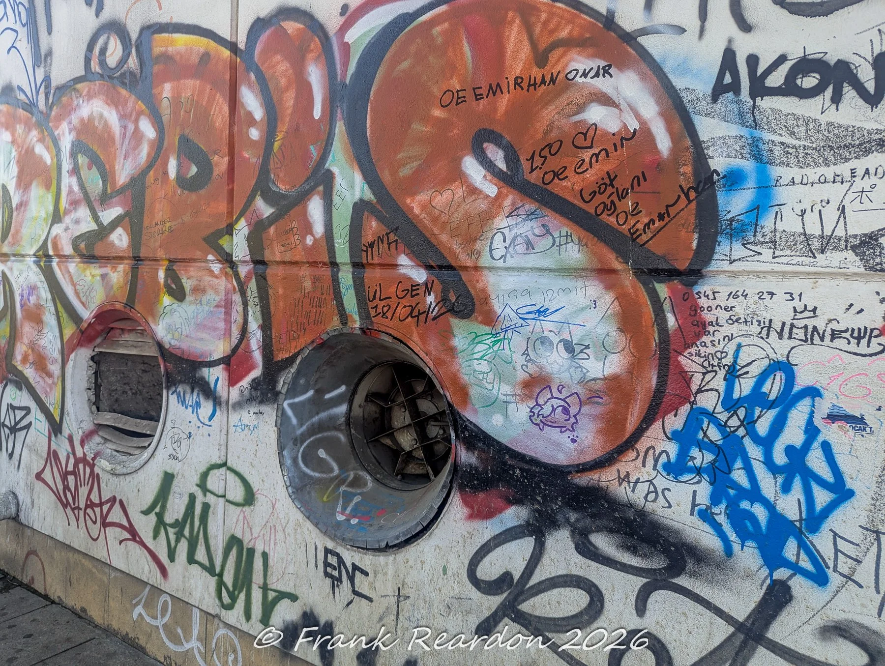 A weathered urban wall featuring large, colorful bubble-letter graffiti tags layered over built-in circular utility ports and vents.