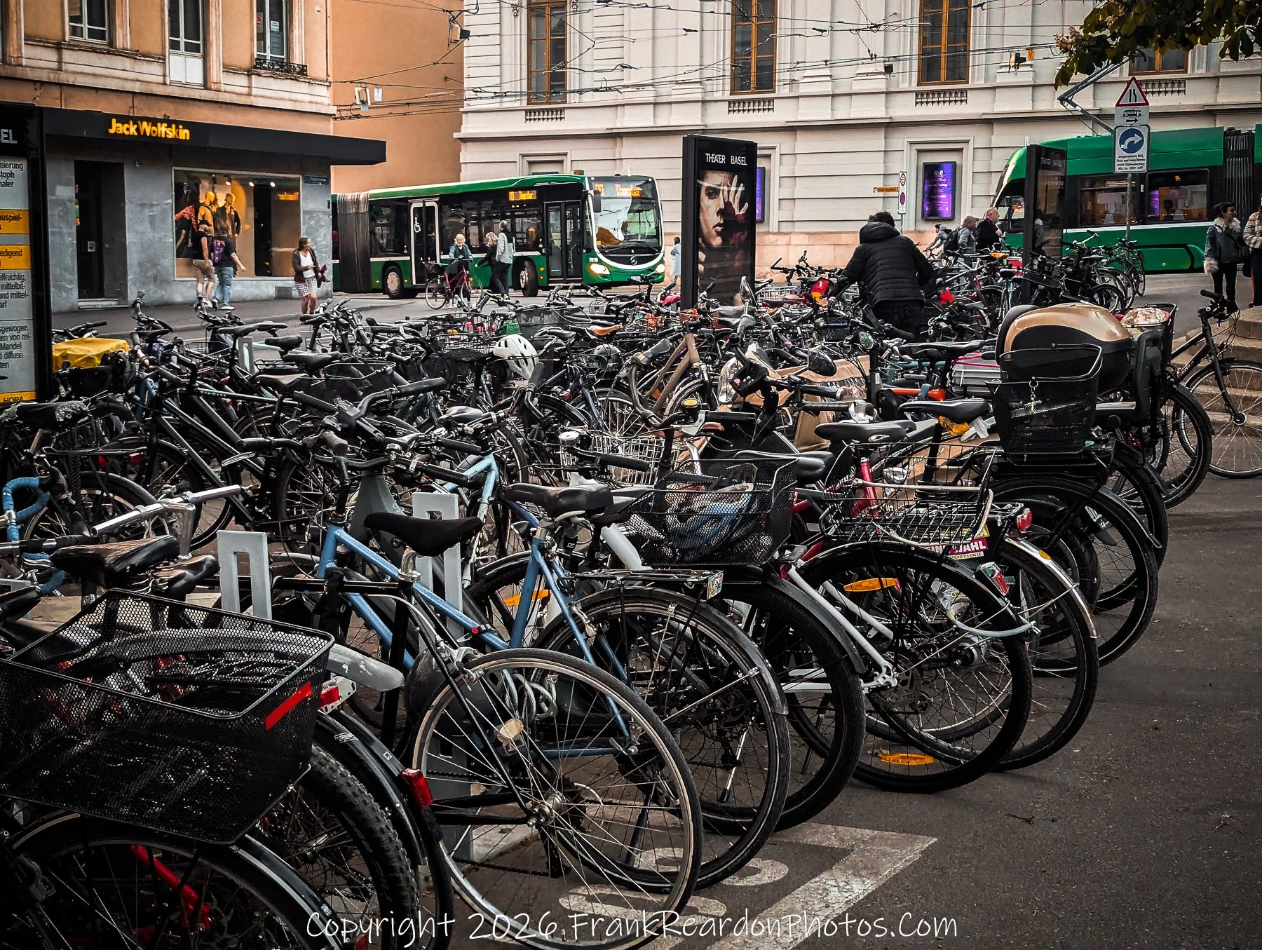 "A Dense Urban Bike Rack" A crowded public bicycle parking area filled with numerous bikes, with green public transport buses visible in the background near a "Jack Wolfskin" storefront.