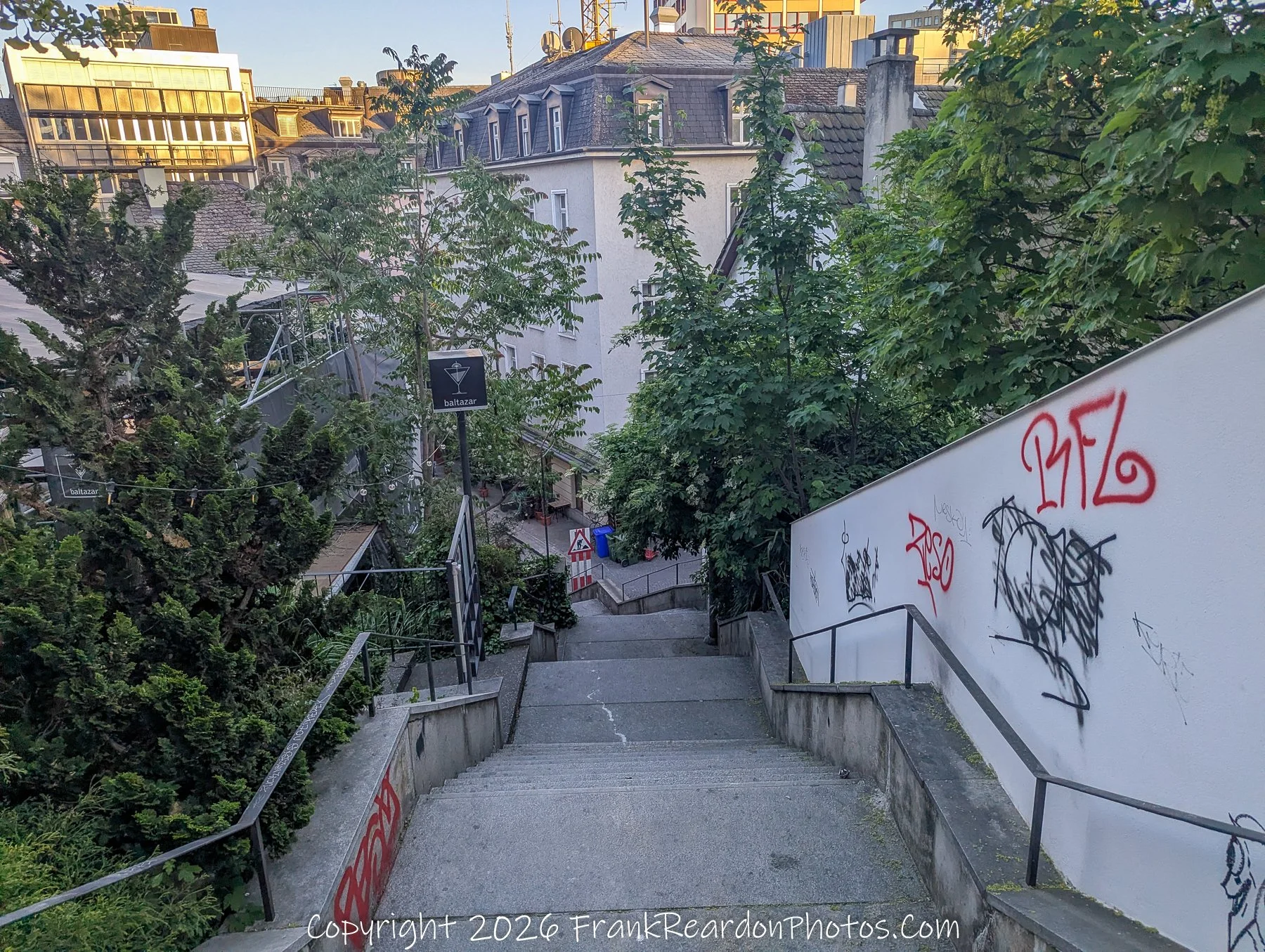 "Stairway to the Hidden Café" A steep set of stone stairs leading down into a leafy urban area, marked by graffiti on a white wall and a sign for "Baltazar" nearby.