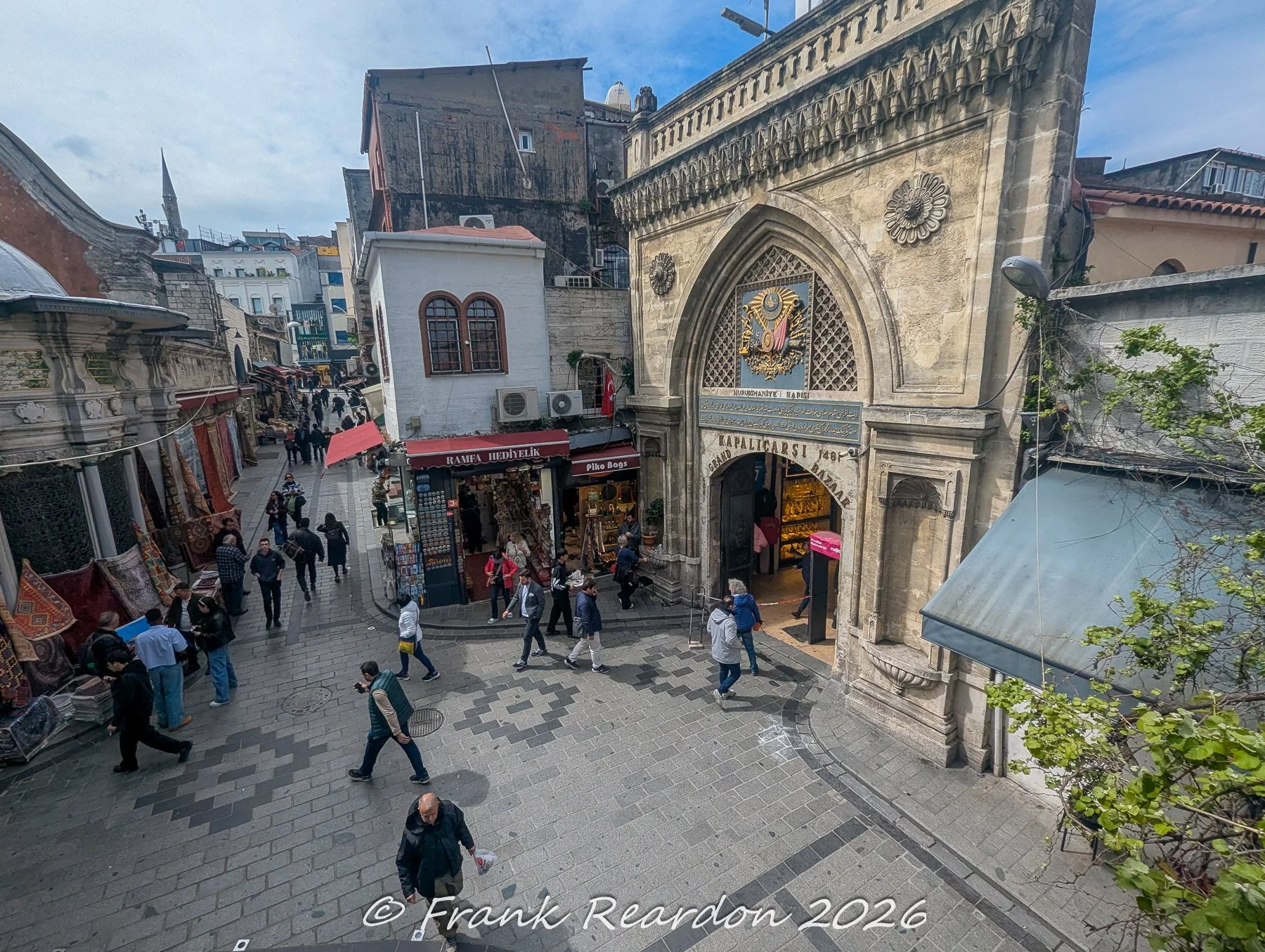 A wide-angle shot looking down at the historic stone entry arch and bustling pedestrians entering the Grand Bazaar (Kapalıçarşı).