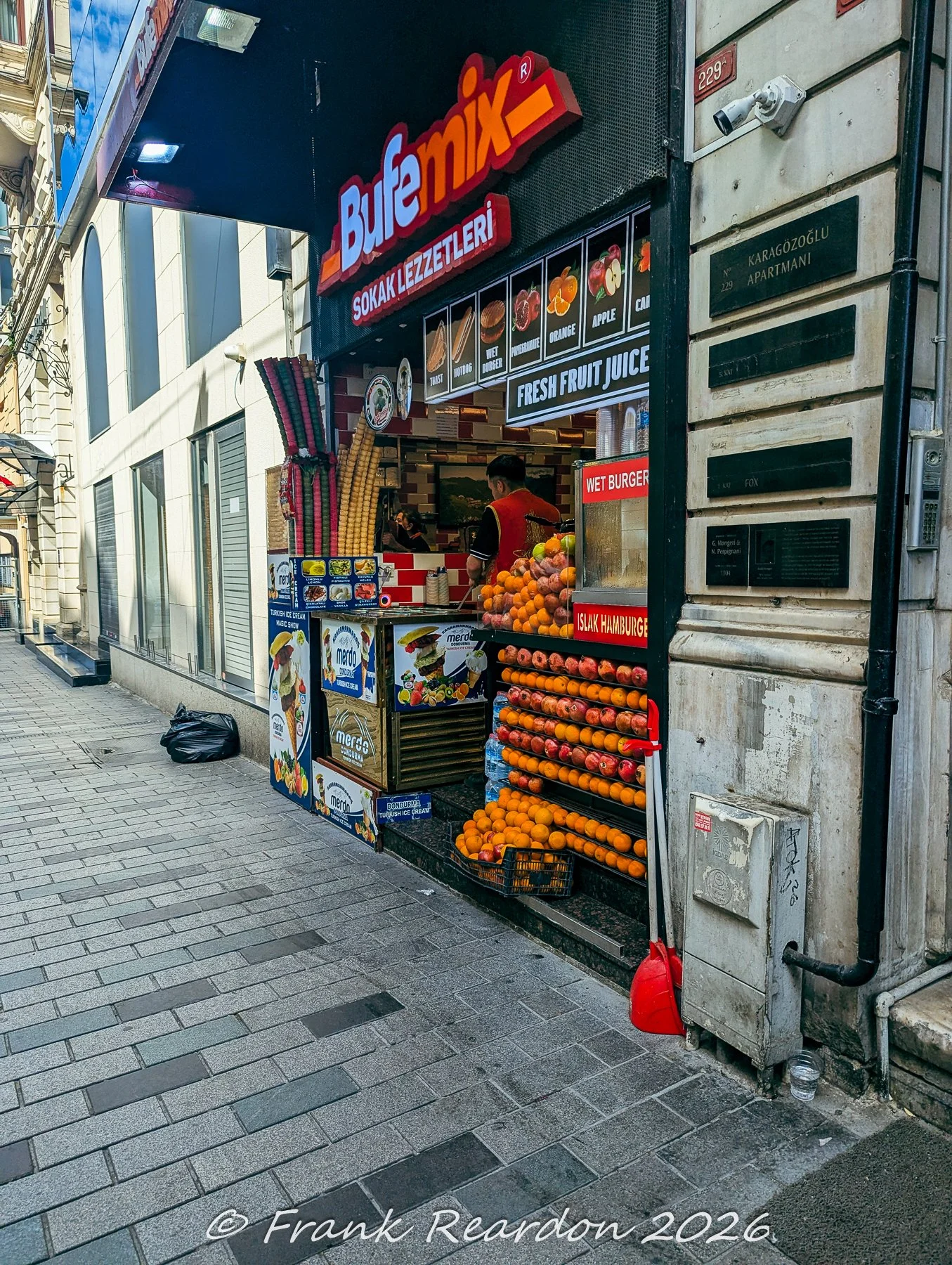 A local street-food stand ("Sokak Lezzetleri") displaying fresh fruit, juice, and snacks next to a doorway labeled Karagözoğlu Apartmanı.