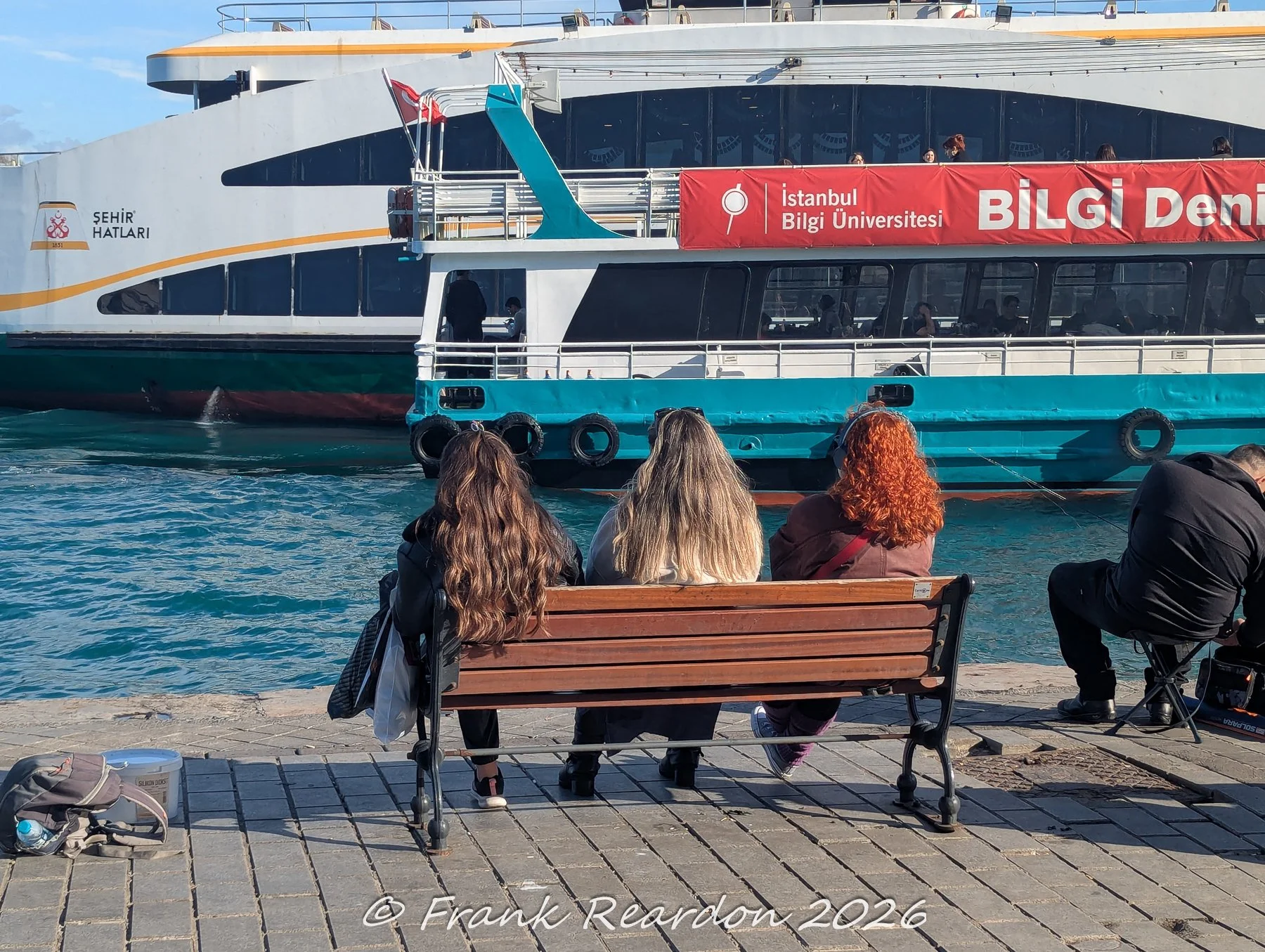 Three people sitting on a wooden bench from behind, looking out at a large municipal ferry (Şehir Hatları) and a Bilgi University shuttle boat docked along the bustling waterfront.