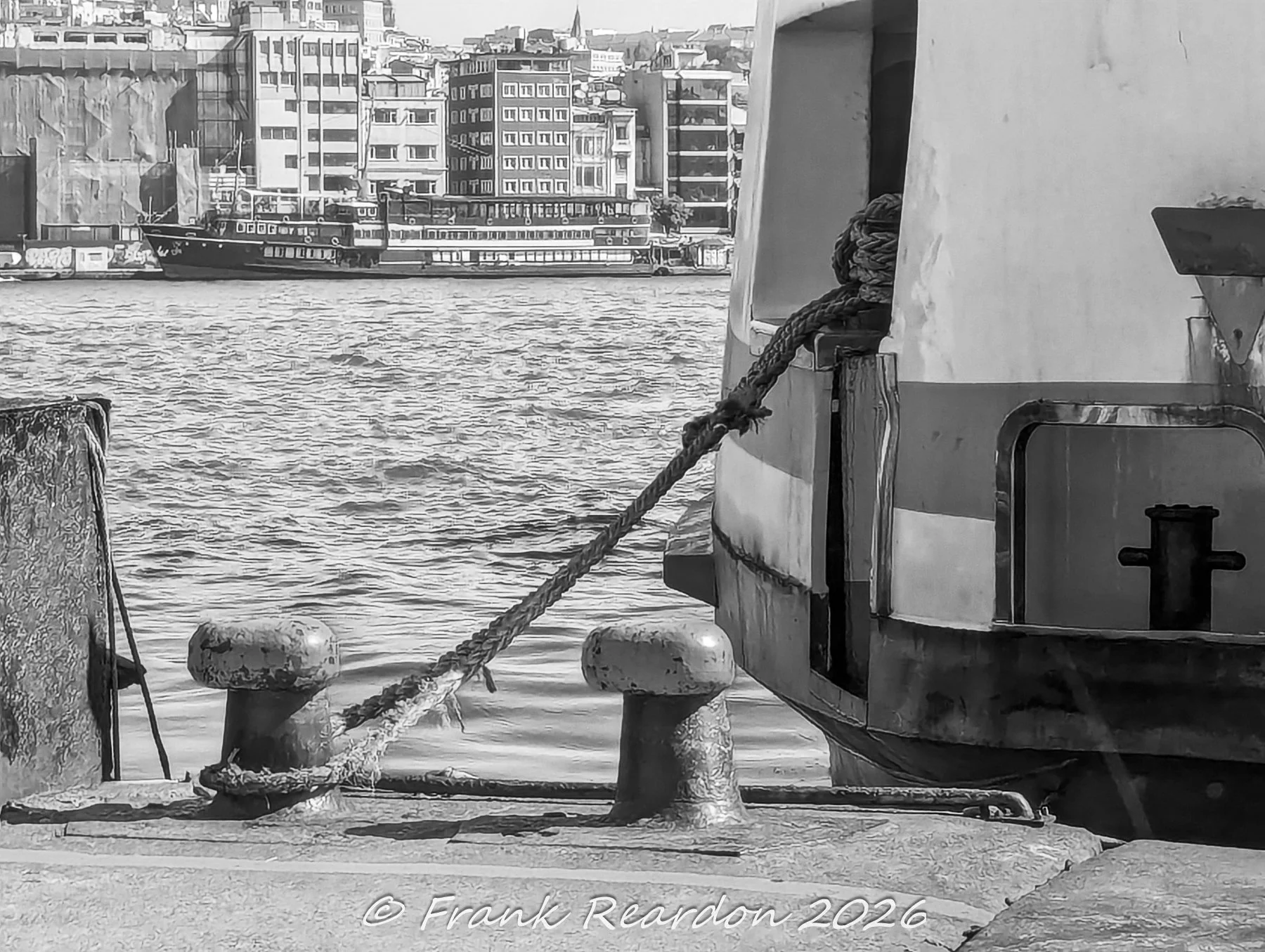 A black-and-white scene capturing a thick mooring rope secured to cast-iron bollards on the stone quay, with a ferry hull and distant Istanbul buildings across the water.